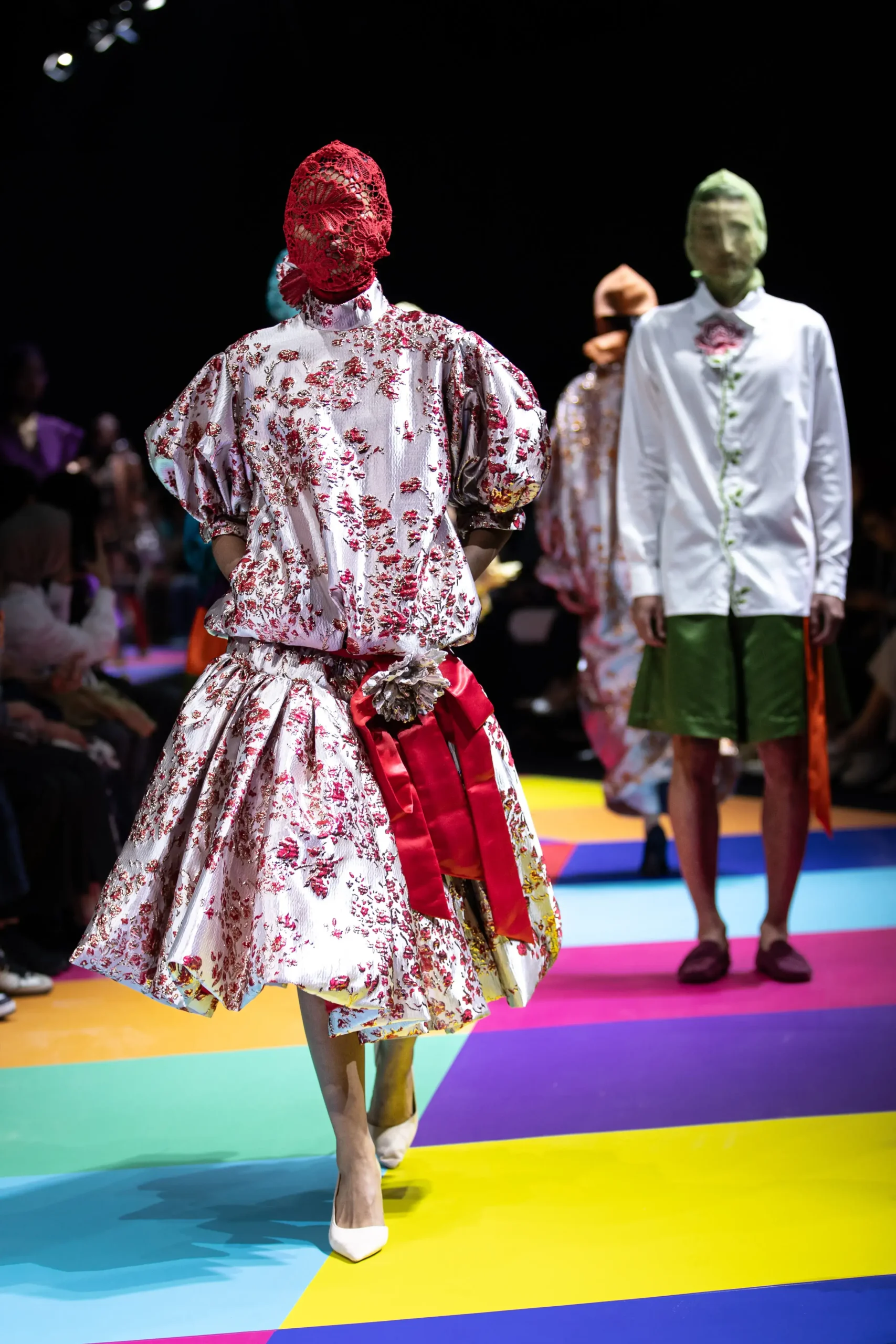Model in a white floral gown with a red headpiece walks a multicolored runway during a fashion show, with other models in the background.