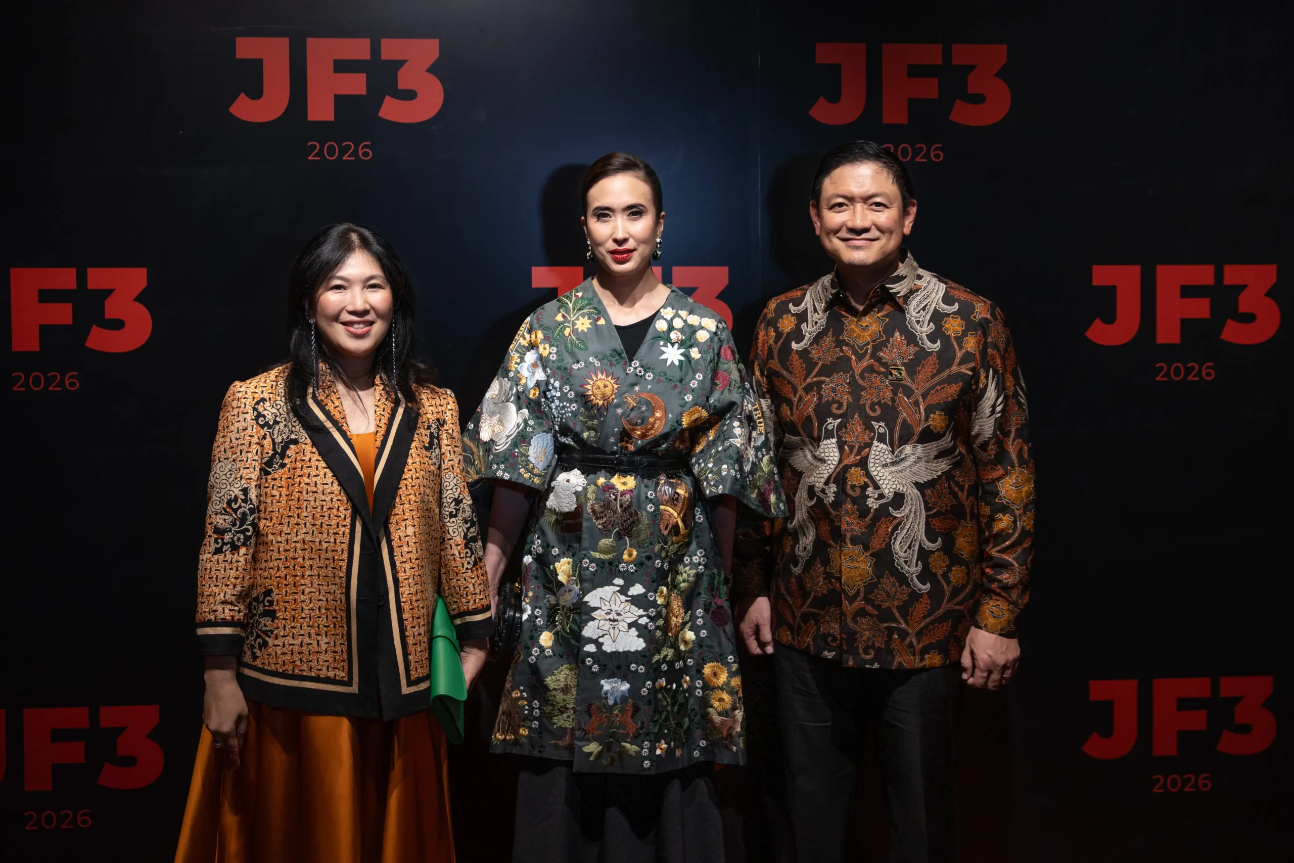 Three people stand against a black backdrop with red JF3 2026 logos, wearing richly patterned, traditional-style outfits.