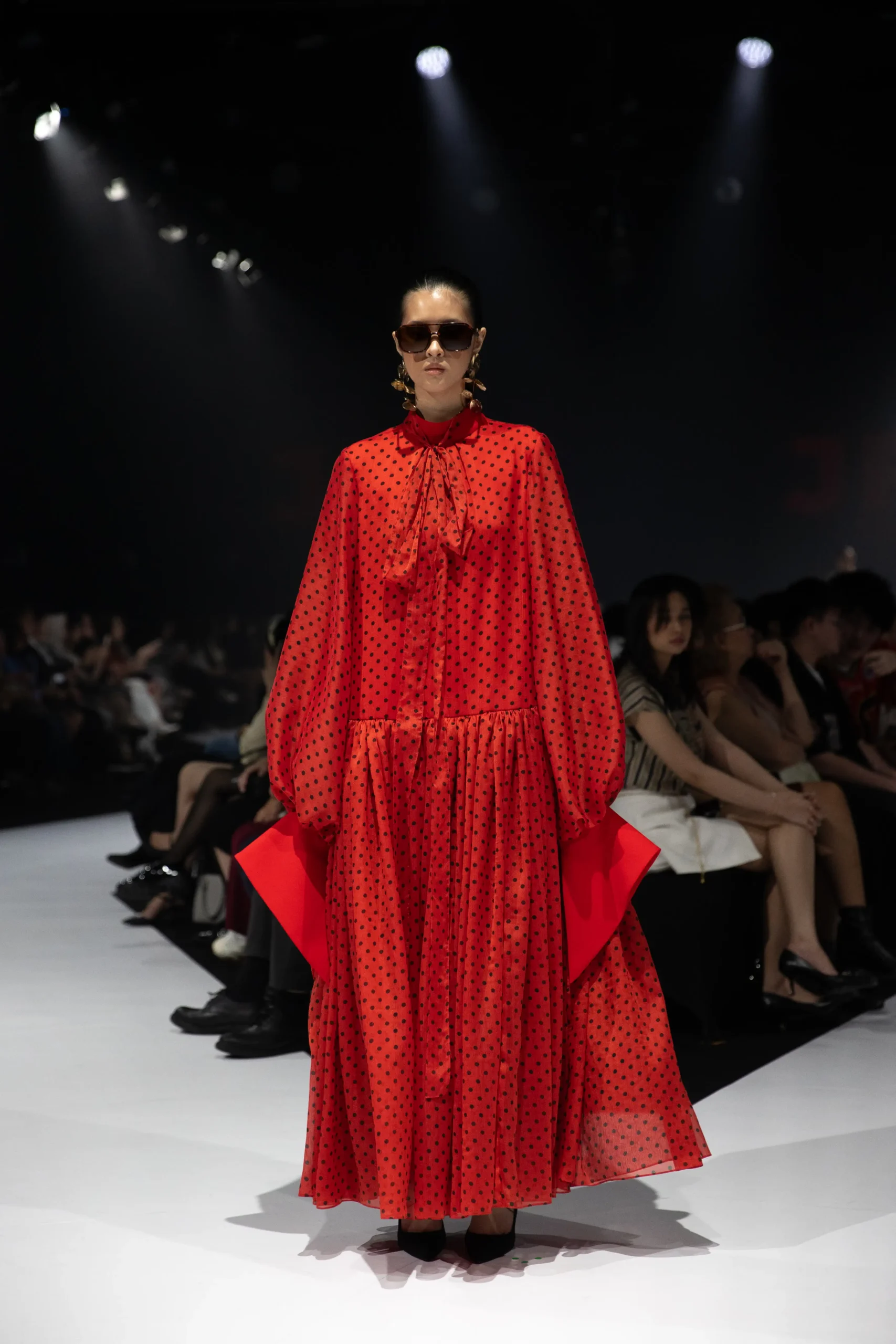 Model on a runway wearing a red polka-dot maxi dress with a bow-tied neckline, oversized sleeves, and sunglasses under spotlights.