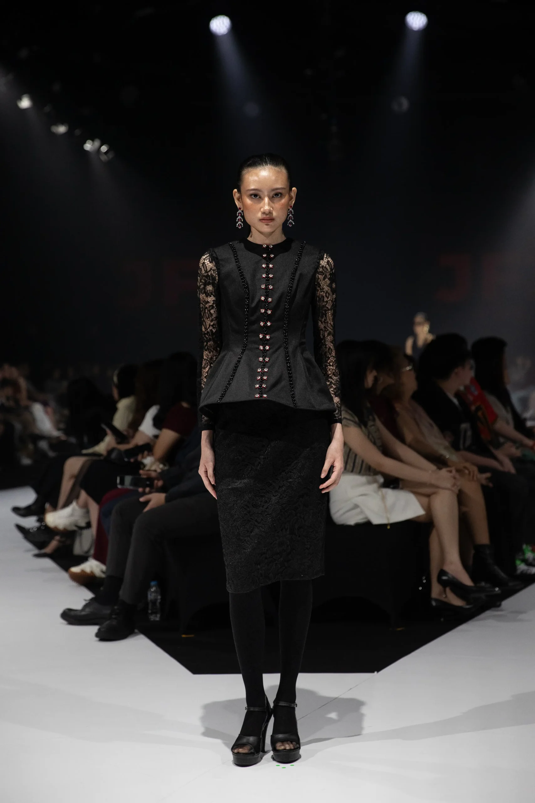 Model walks a black lace-sleeved dress down the runway under spotlights, преступно black attire with red button detailing.