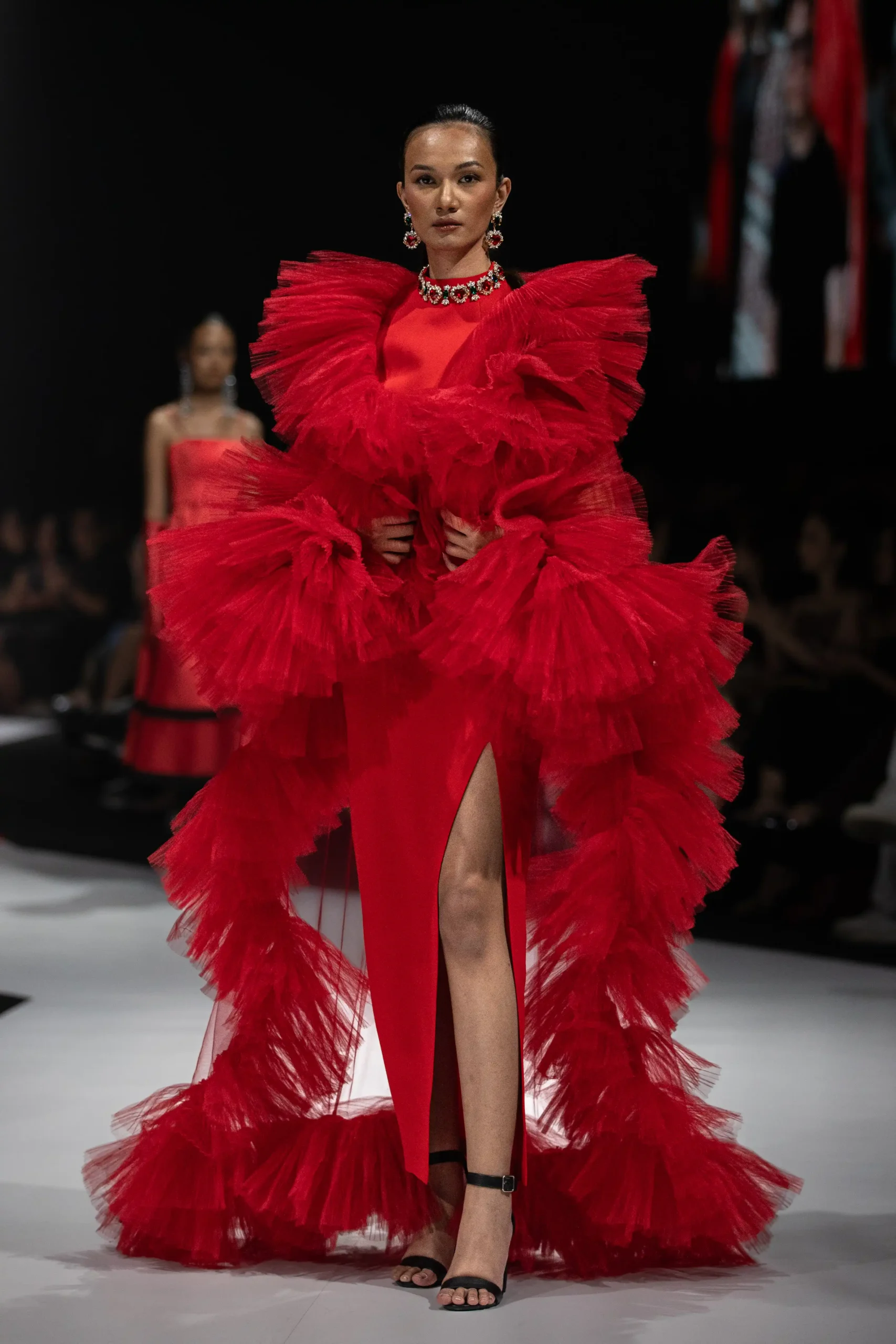 Runway model wearing a bold red, ruffled gown with voluminous tulle and statement jewelry on the catwalk.