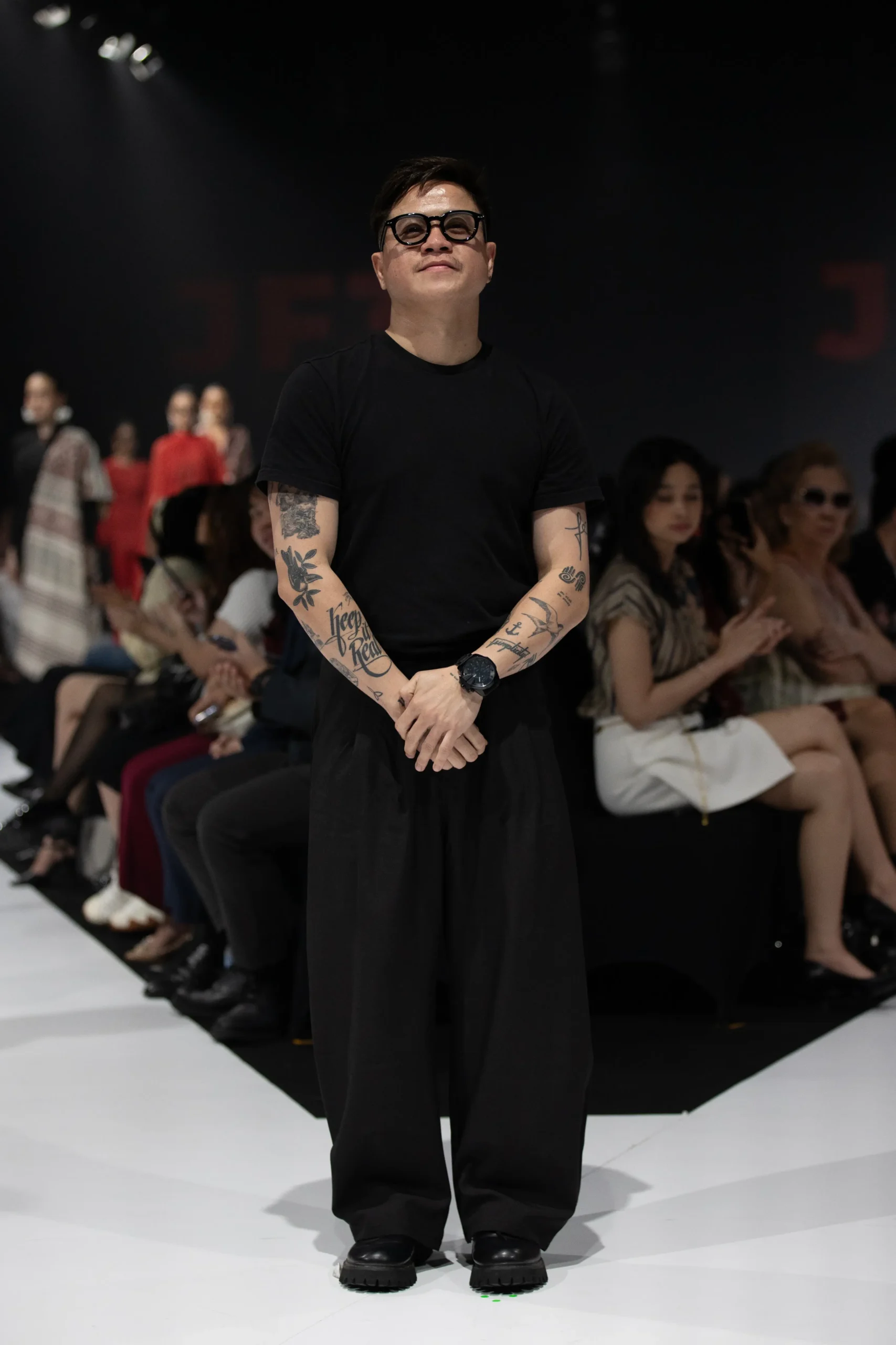 Male fashion model in a black T-shirt and wide black pants, with tattoos on both arms, standing on a runway runway view.