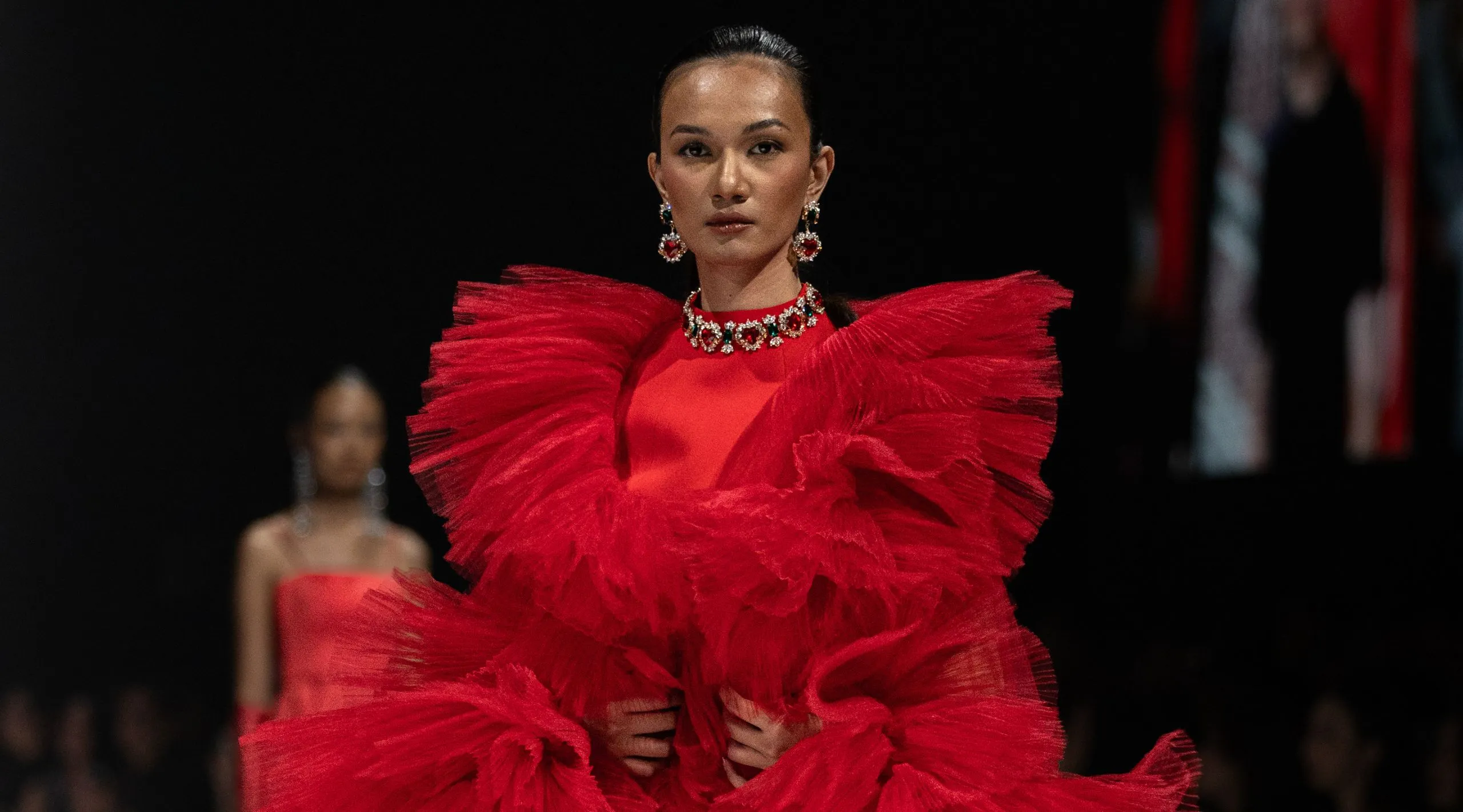 Model on a runway wearing a red, voluminous ruffled gown with a jeweled necklace and earrings against a dark background.