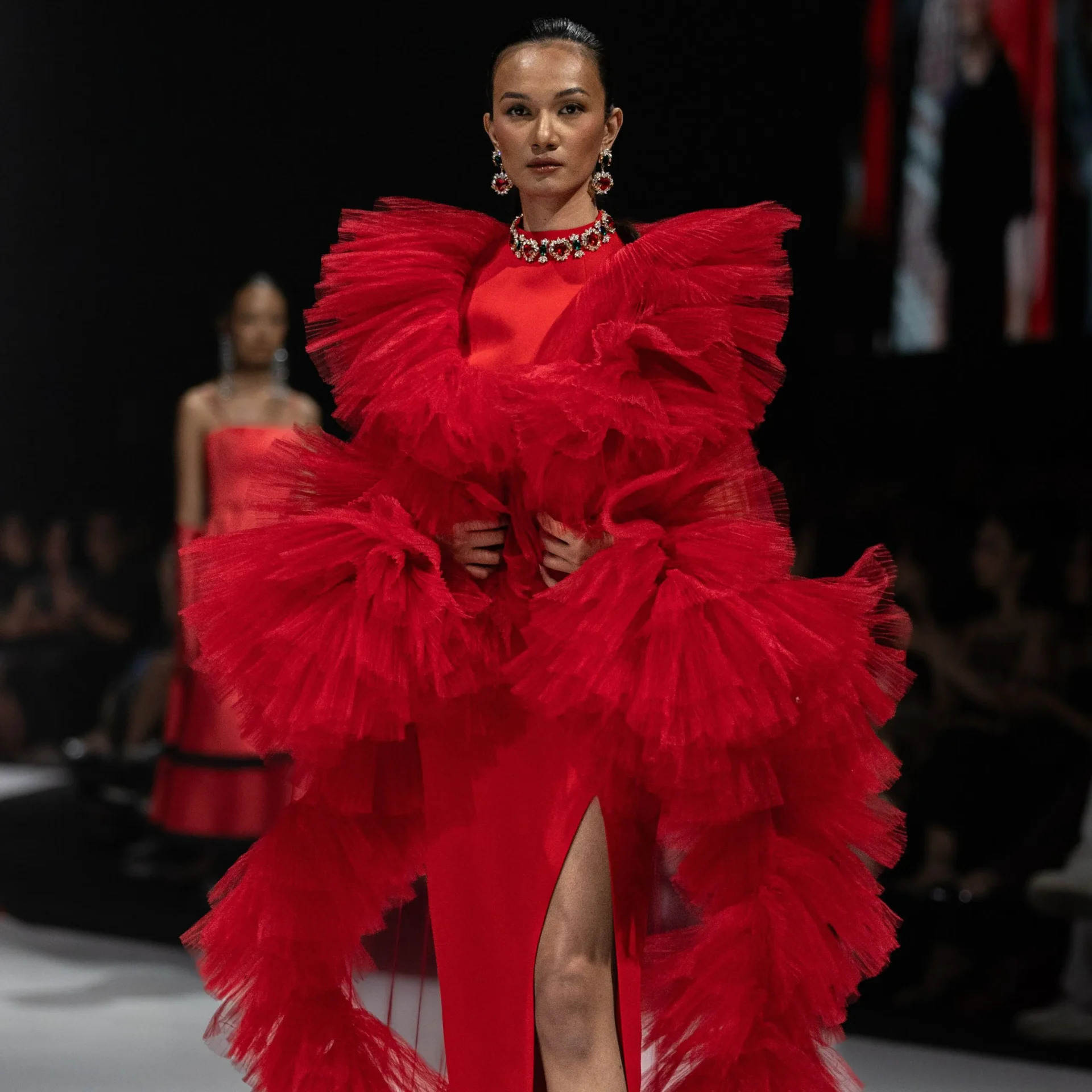 Model wearing a dramatic red ruffled gown and statement jewelry on a fashion runway.