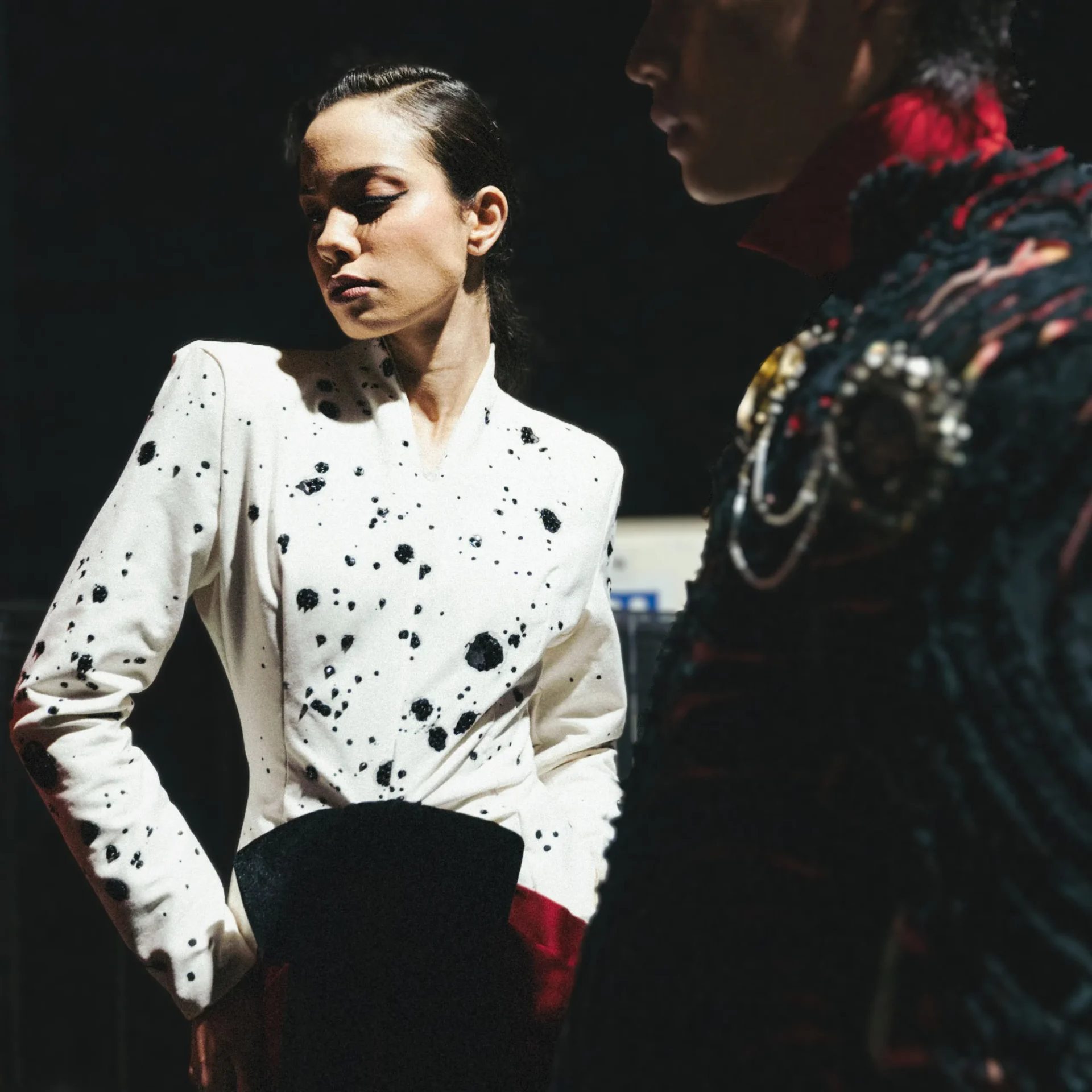 Woman in a white, black-splattered top stands in a dark setting, next to a person wearing a colorful patterned outfit.