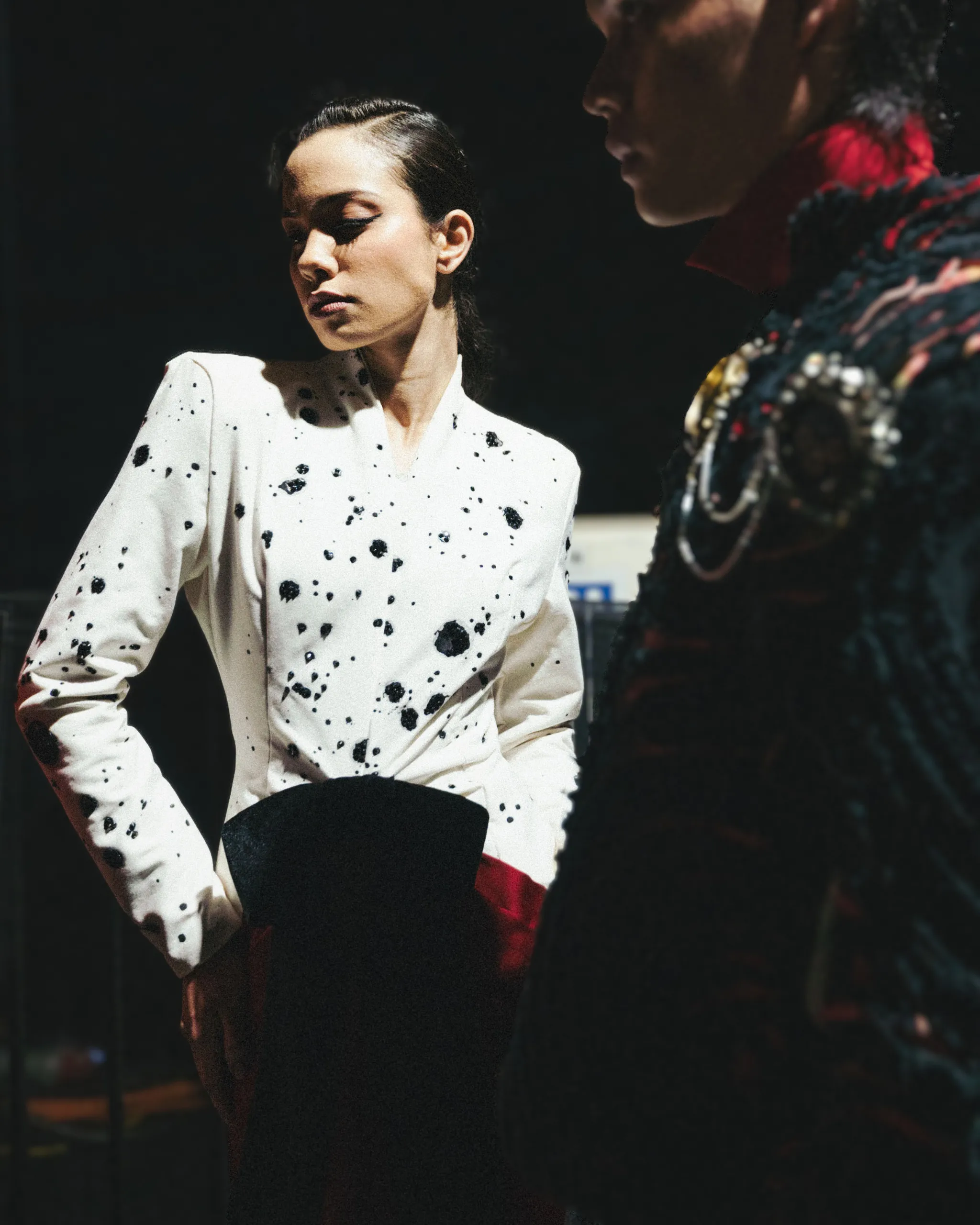 Woman in a white blouse with black splatters, eyes closed, standing beside a second person in a dark patterned outfit in low light.