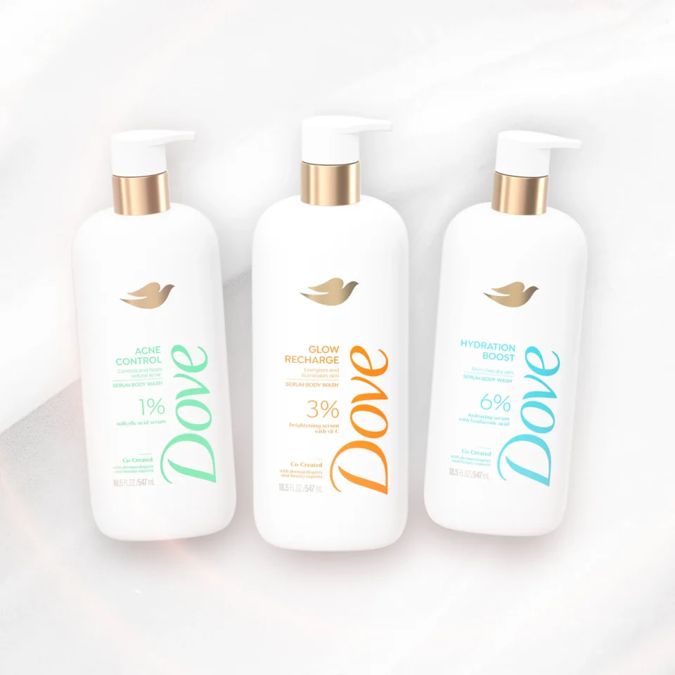 Three white pump bottles with gold collars arranged in a row on a light marble surface, showcasing skincare product display.