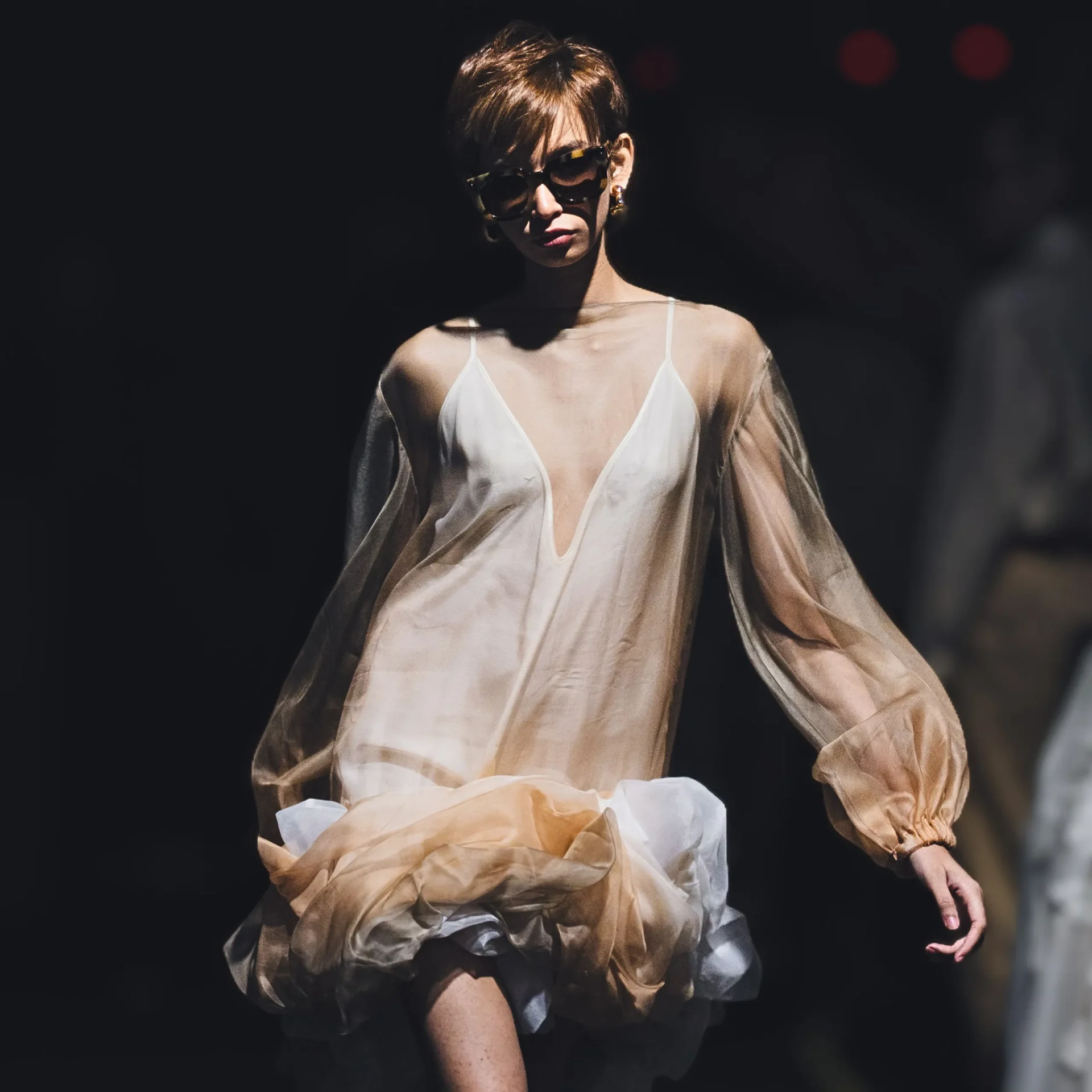 Model on a dark runway wearing a sheer beige outer layer, white dress, and large sunglasses.