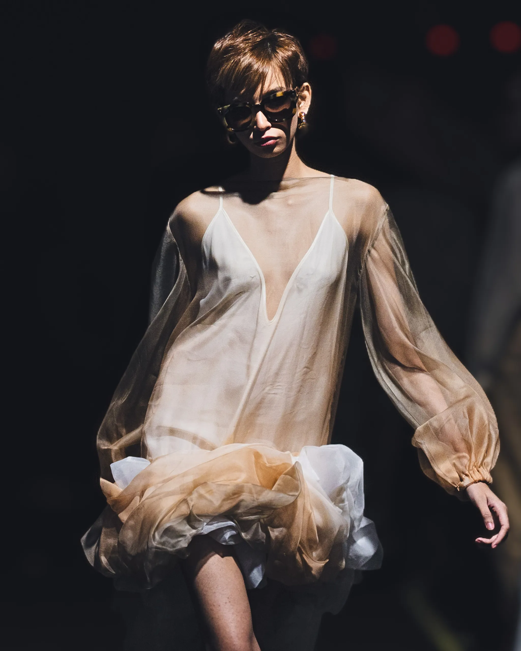 Fashion model on a dark runway wearing a sheer beige gown and oversized sunglasses, spotlight on her.