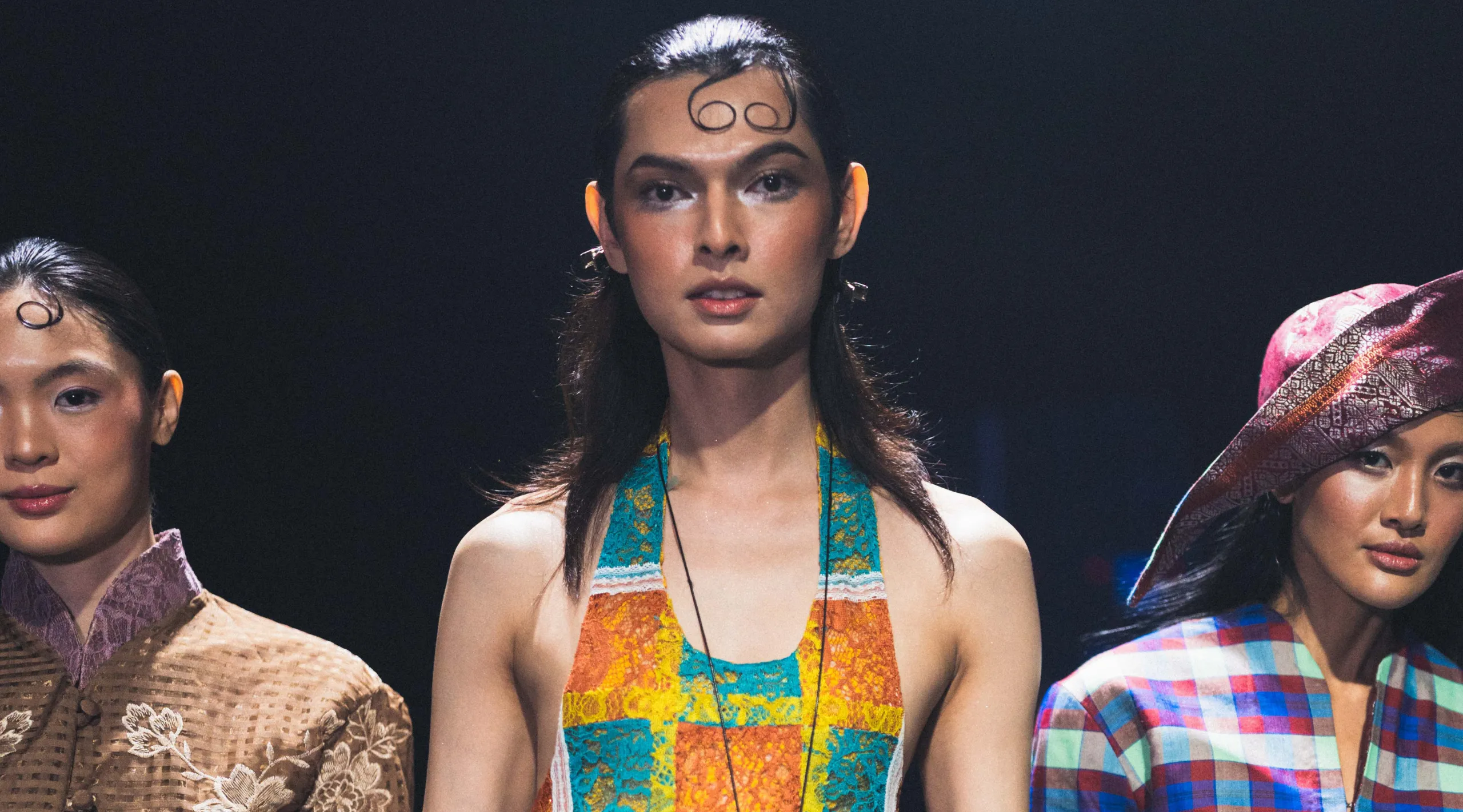 Center-stage fashion model in a colorful turquoise, orange, and yellow halter top, flanked by two others in patterned outfits on a dark runway backdrop.