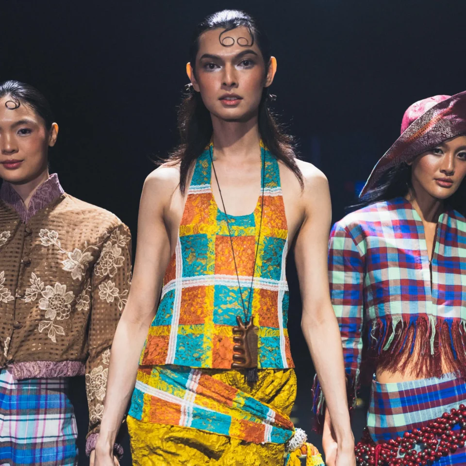 Three models on a fashion runway wearing colorful patchwork dresses, center foreground.