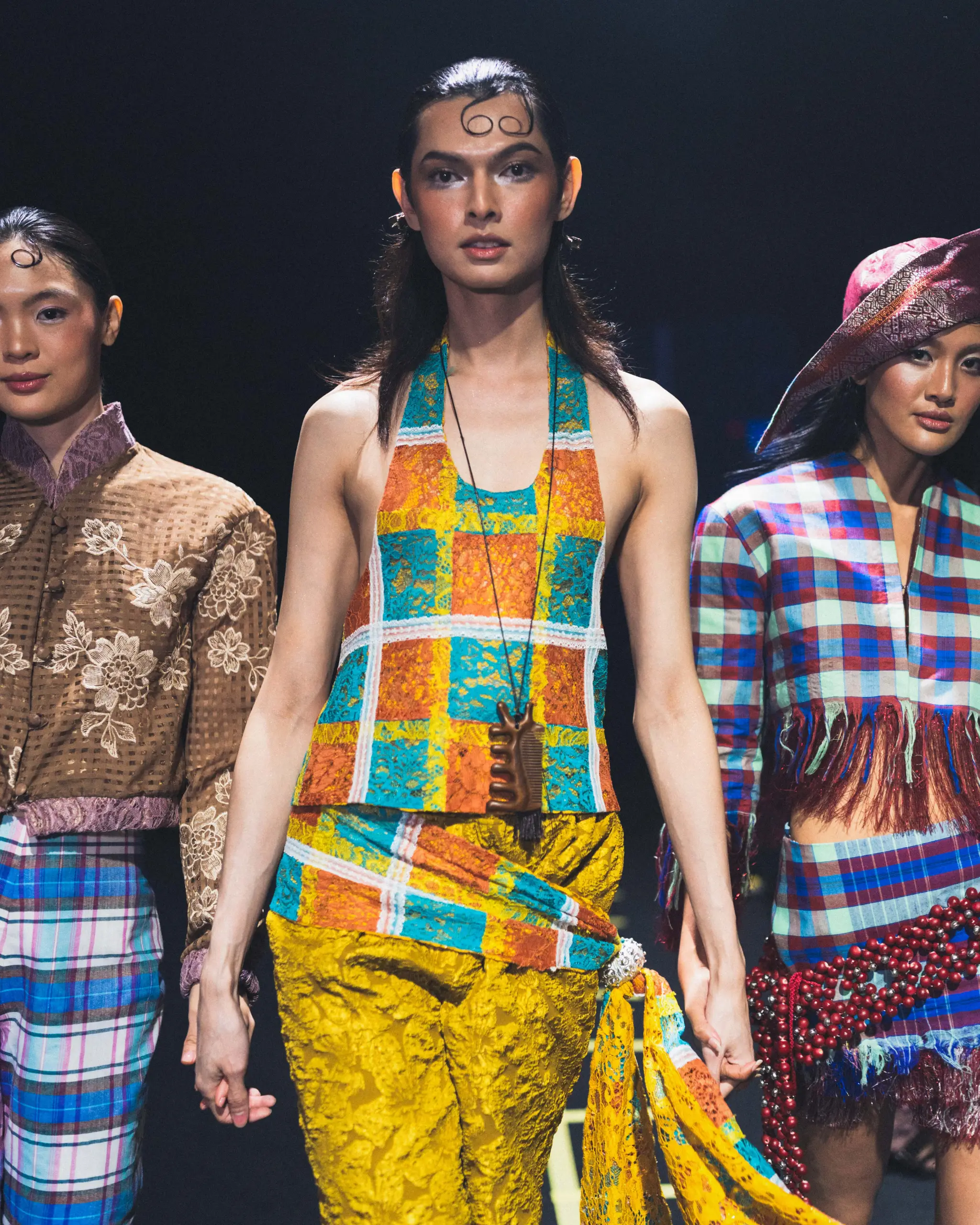 Three fashion models stand on a runway in bold, colorful outfits; center model wears a yellow, turquoise, and orange lace dress with a long necklace.