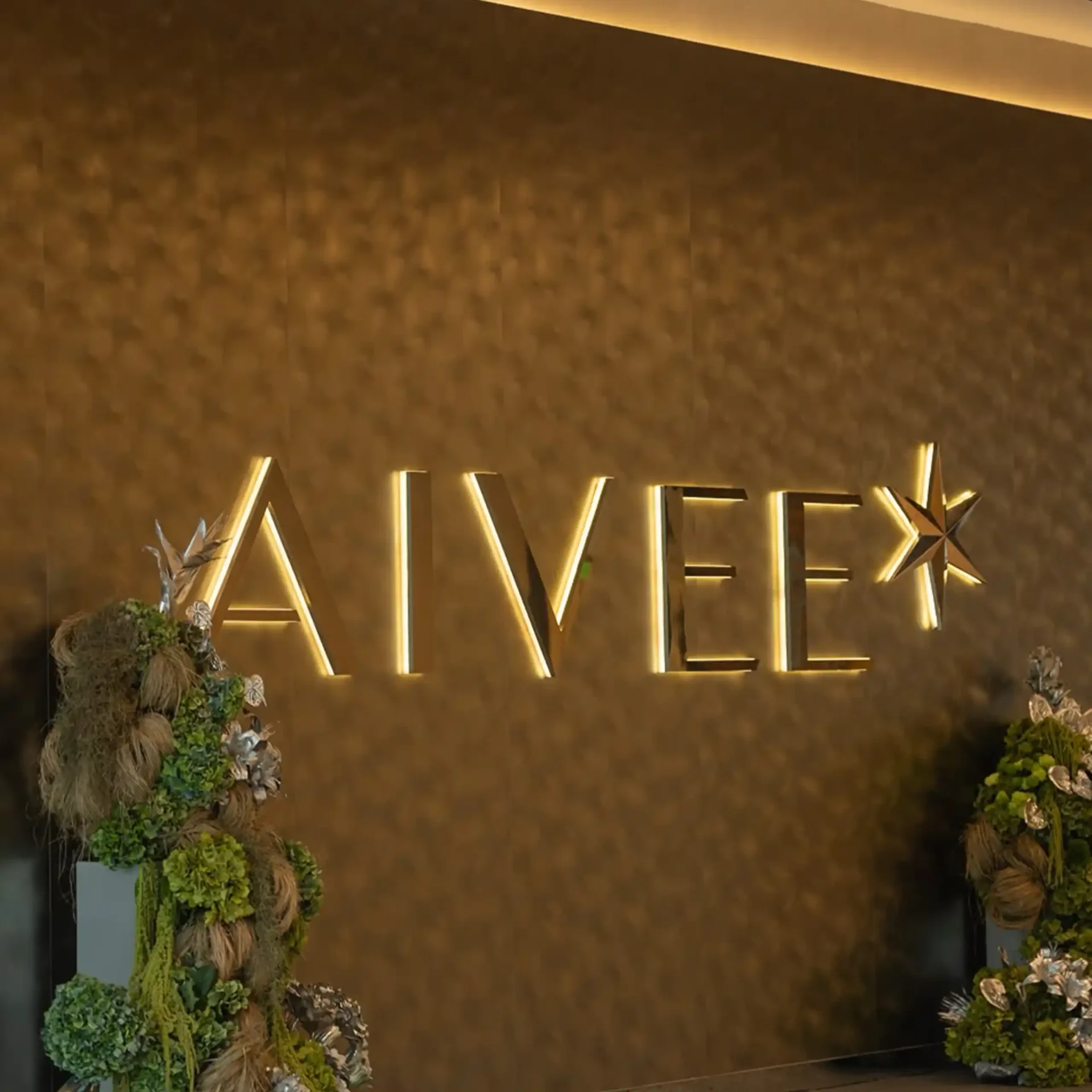 Gold-toned wall backdrop with illuminated geometric lines and lush floral arrangements on both sides.