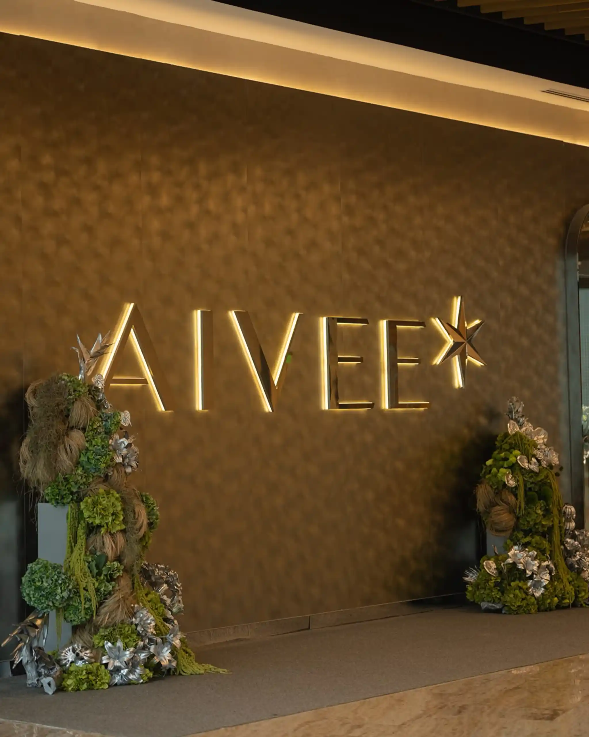 Indoor wall with large, illuminated gold letters forming a logo, flanked by decorative green and metallic floral arrangements.