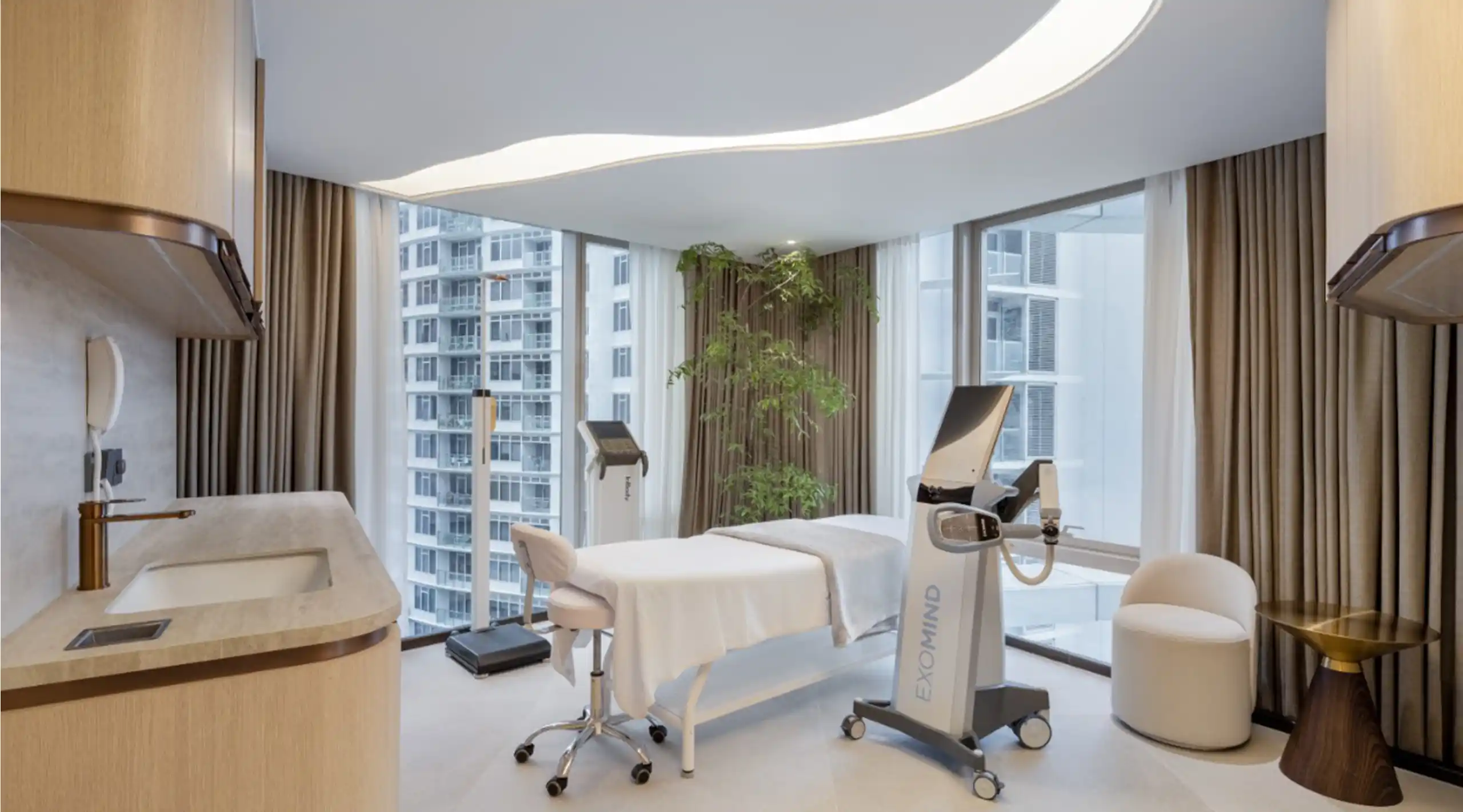 Luxurious treatment room with a white massage bed, a nearby exercise machine, and large windows overlooking the city cityscape.]','