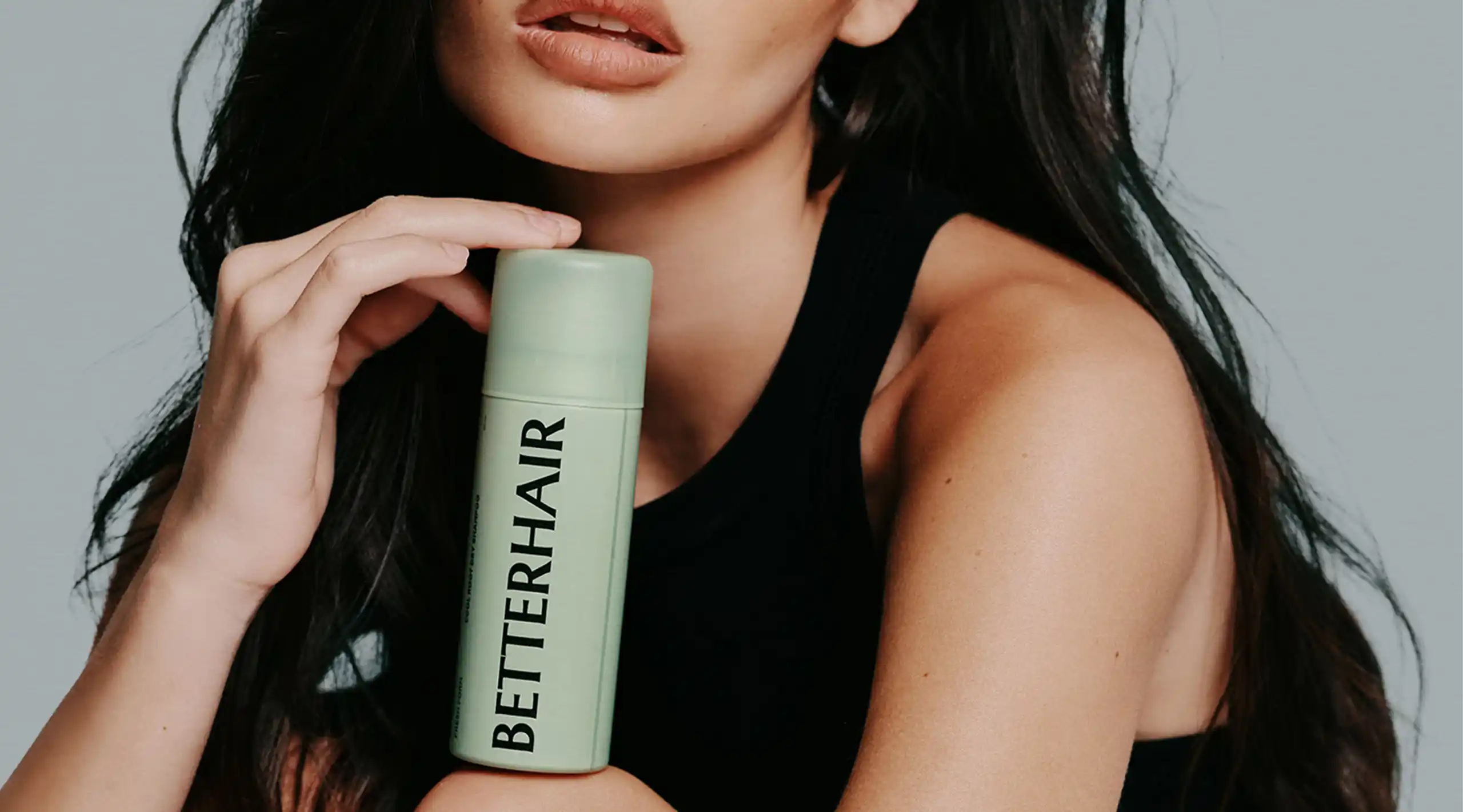 Close-up of a woman with dark hair holding a mint-green skincare bottle against a gray background.