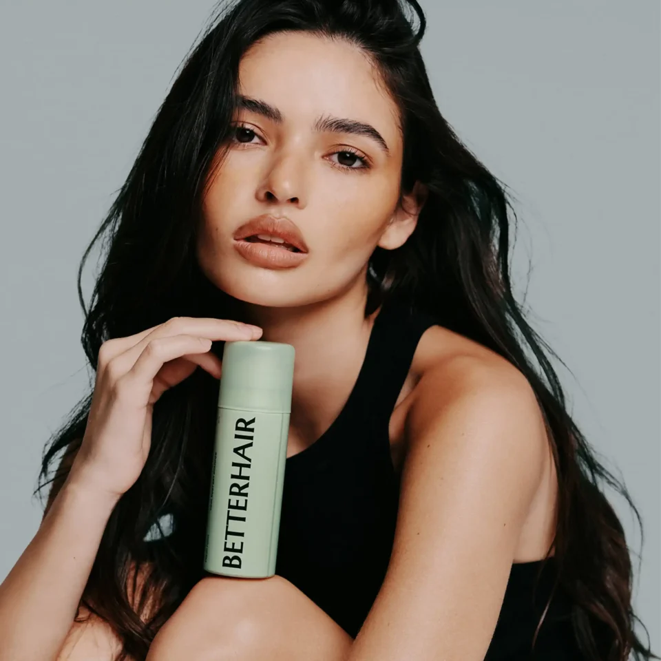 Close-up of a woman with long dark hair holding a light green bottle against a gray background.