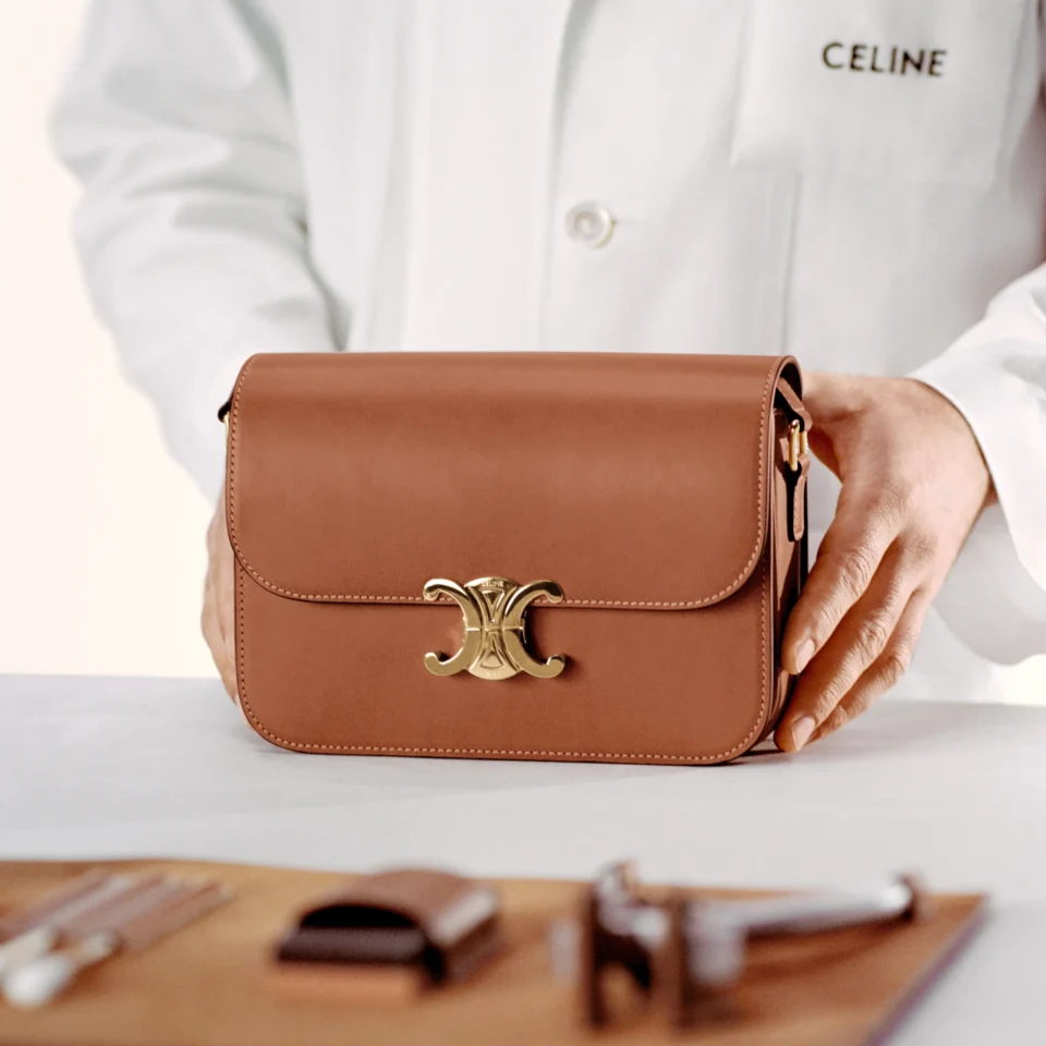 Close-up of a tan leather handbag being held by a person in a white coat over a light background.