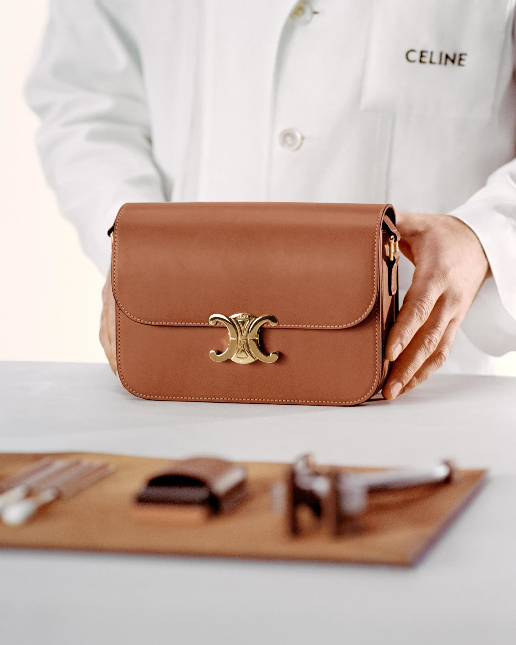Brown leather handbag with gold hardware on a white surface, as a person in a white coat adjusts it in the background.
