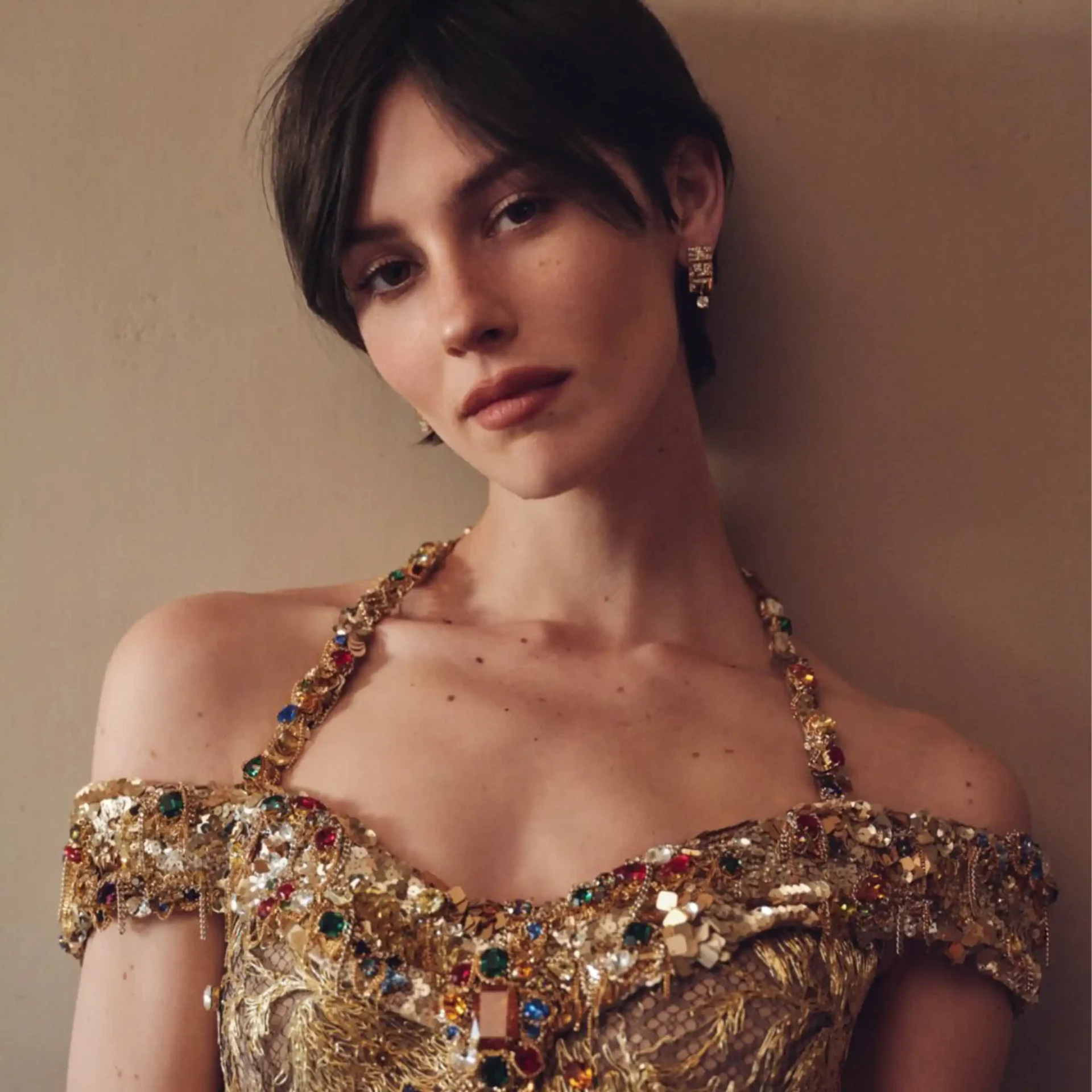 Close-up of a person wearing a gold, jewel-encrusted necklace and matching earrings, with a neutral background.