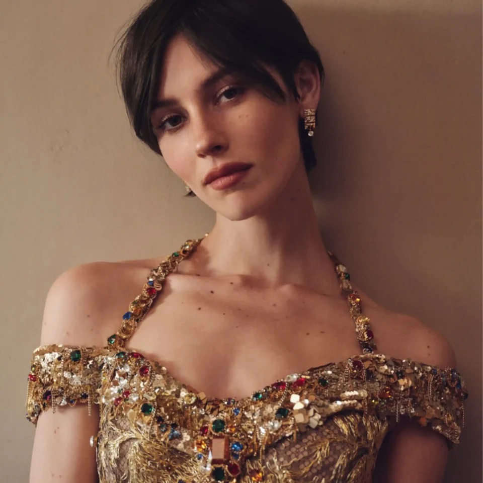 Close-up of a person wearing a gold, jewel-encrusted necklace and matching earrings, with a neutral background.