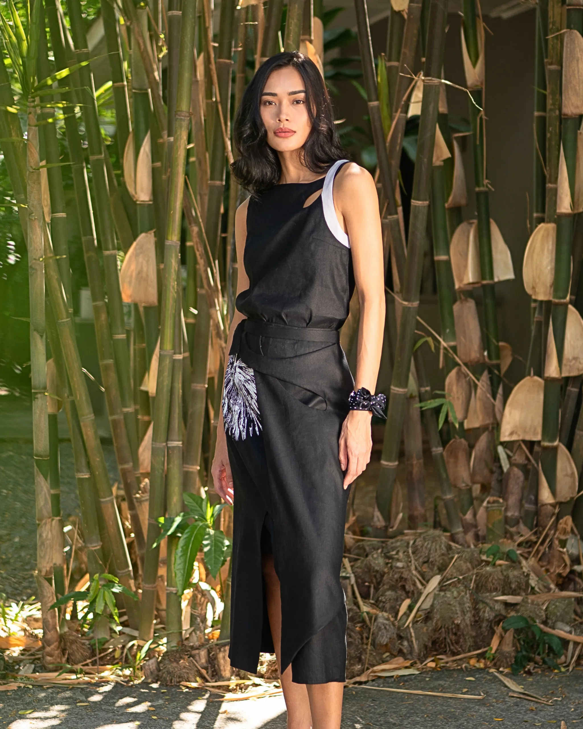 Woman in a black sleeveless dress standing among tall bamboo stalks, posing outdoors in sunlight.
