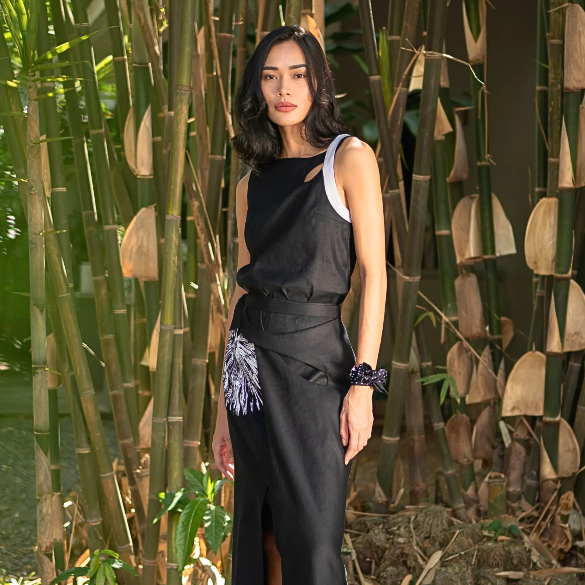 Woman in a black sleeveless dress stands among tall bamboo stalks, facing the camera.