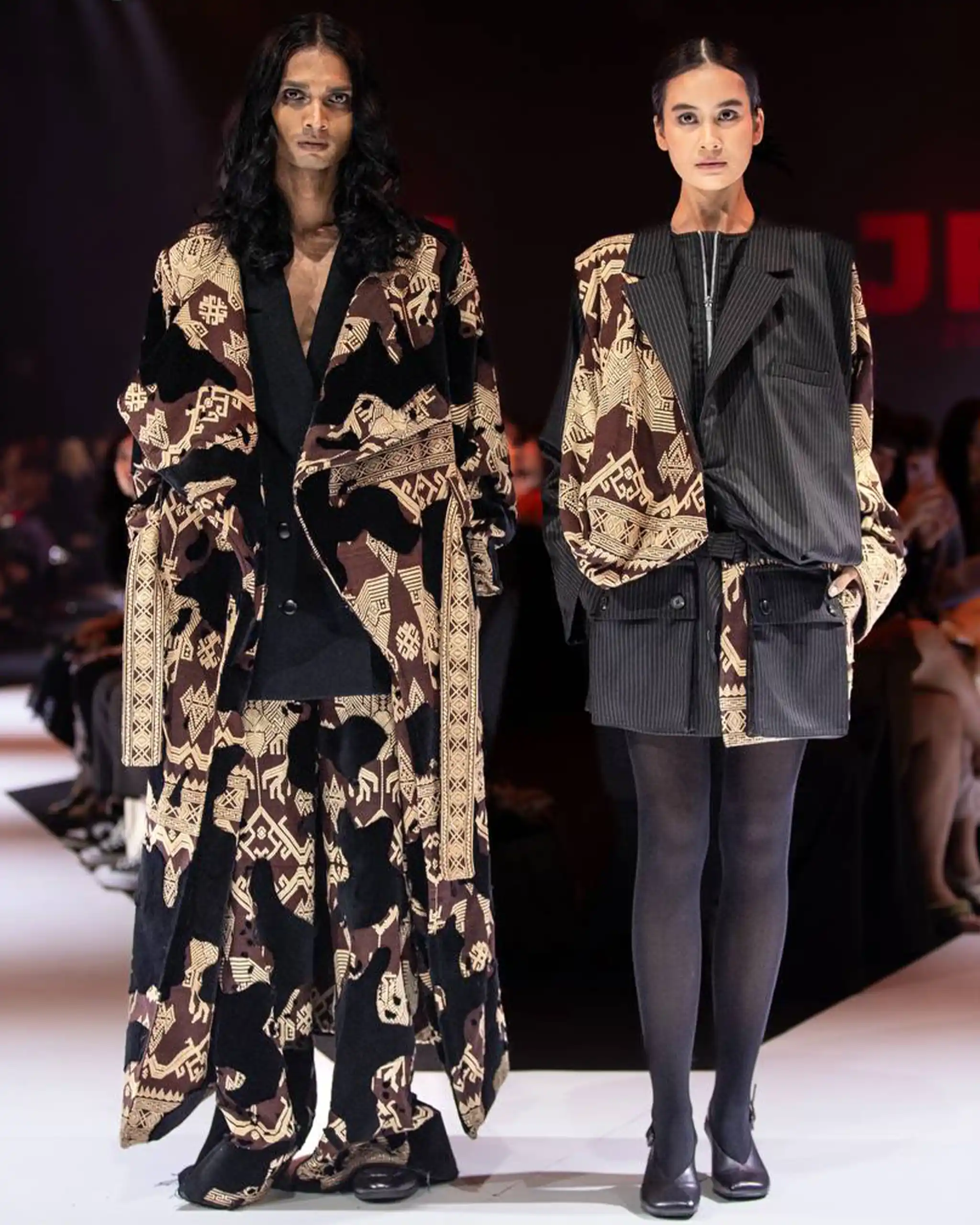 Two models walk a fashion-show runway wearing oversized coats with bold brown geometric patterns.