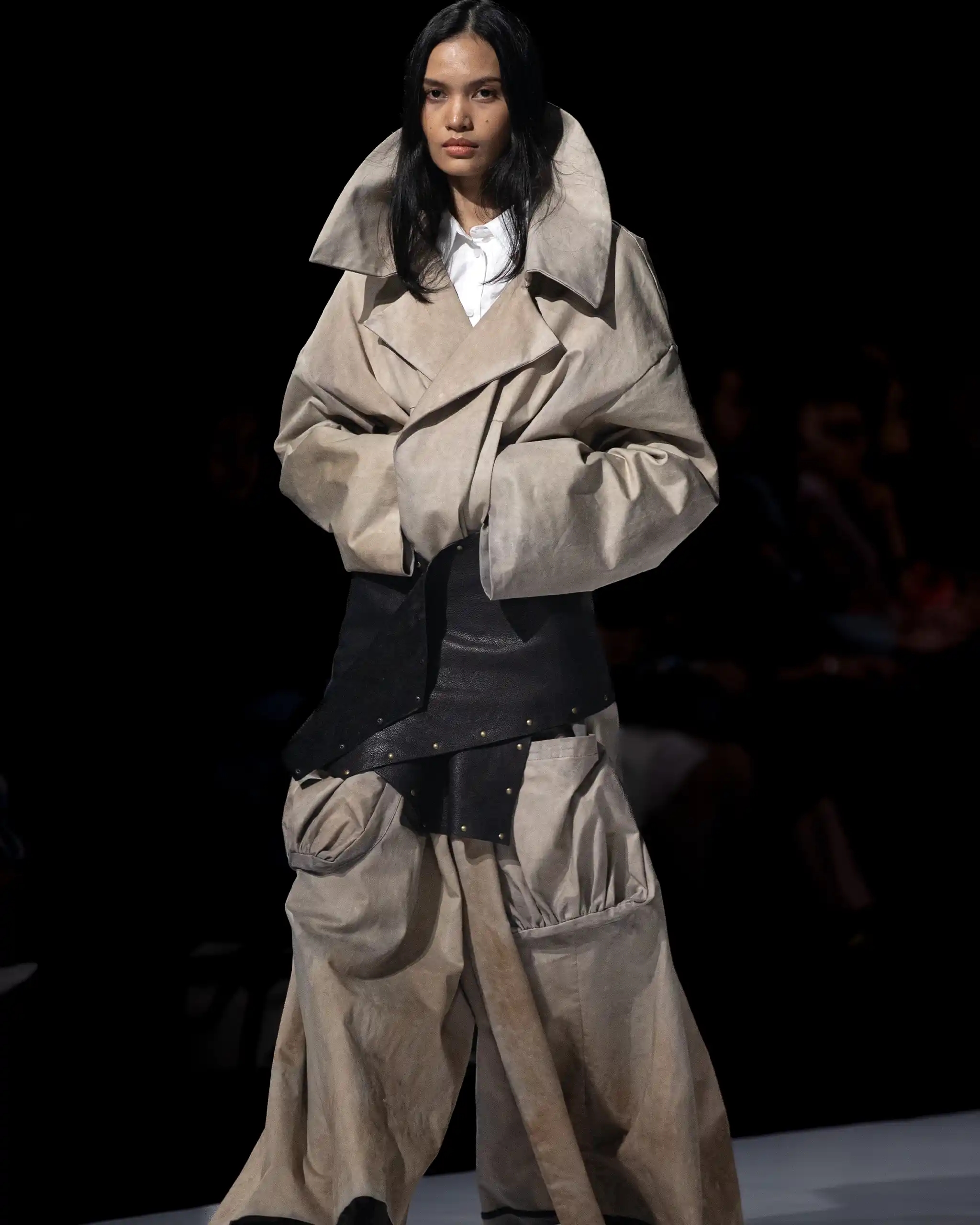 Model walking a runway in a large beige trench coat over a white shirt, against a black background