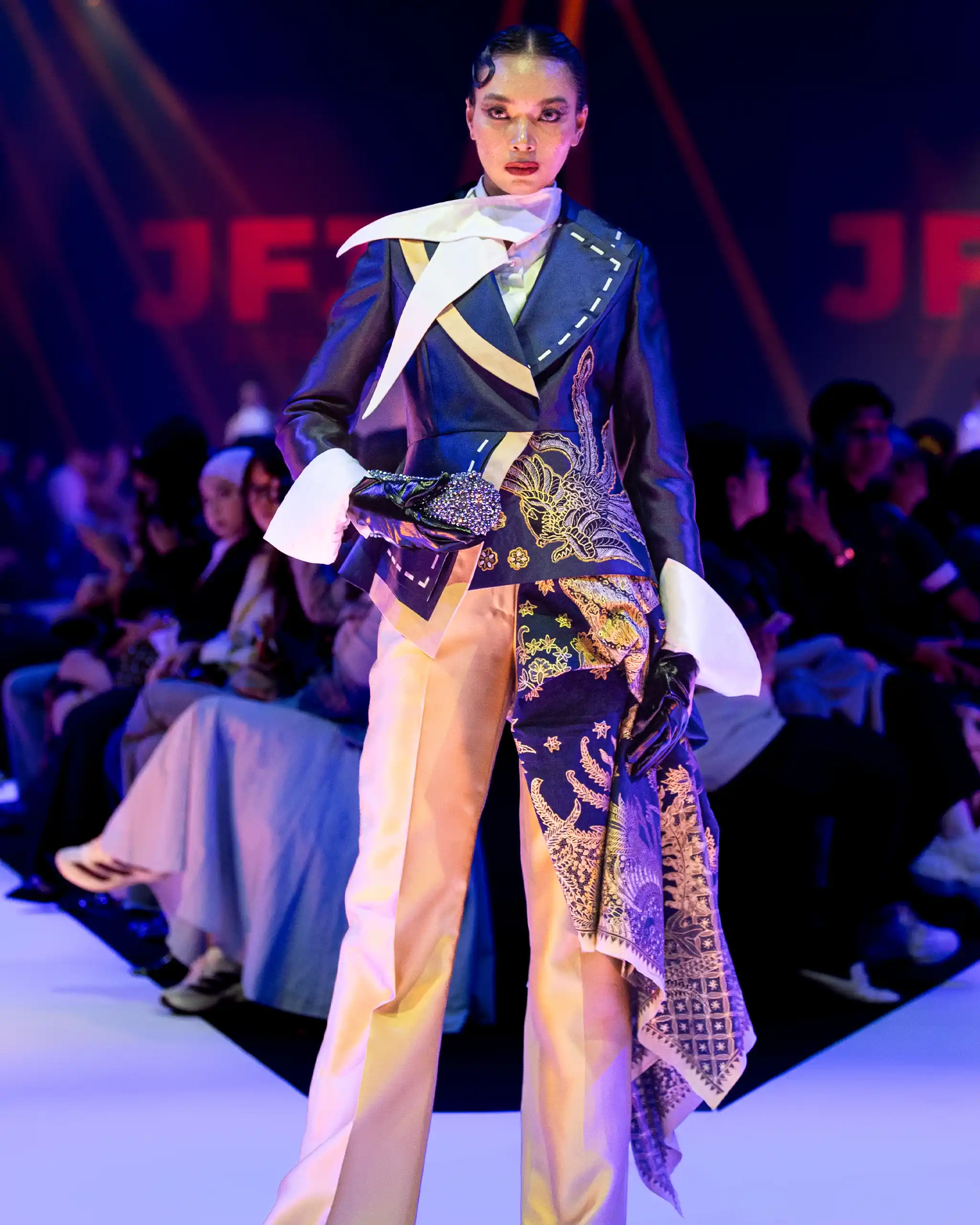Model on a runway wearing a blue and gold patterned suit with a white scarf, spotlighted among a seated audience.