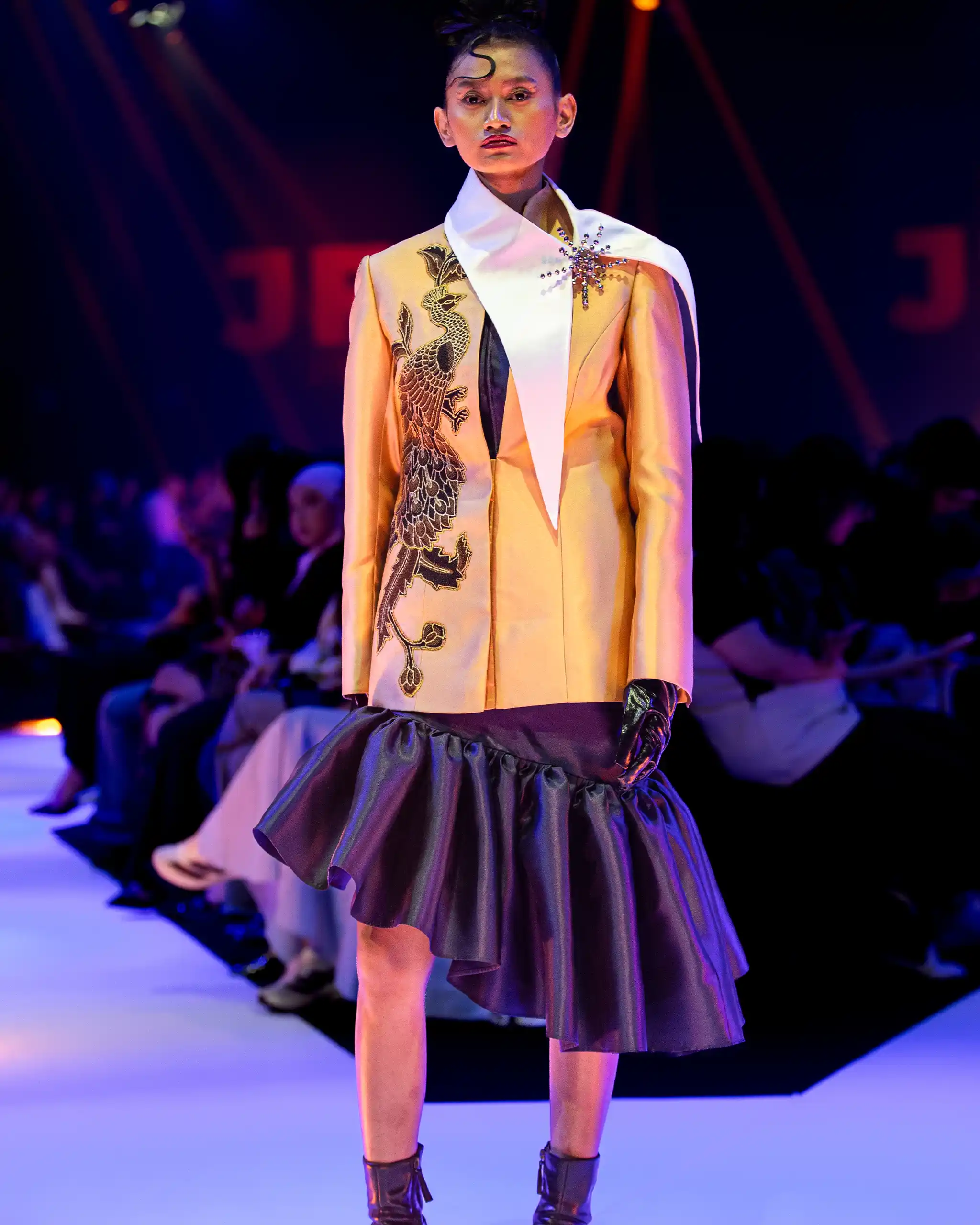 Runway model wearing a gold blazer with dragon embroidery, a white scarf over the shoulder, and a black ruffled skirt under blue stage lighting.