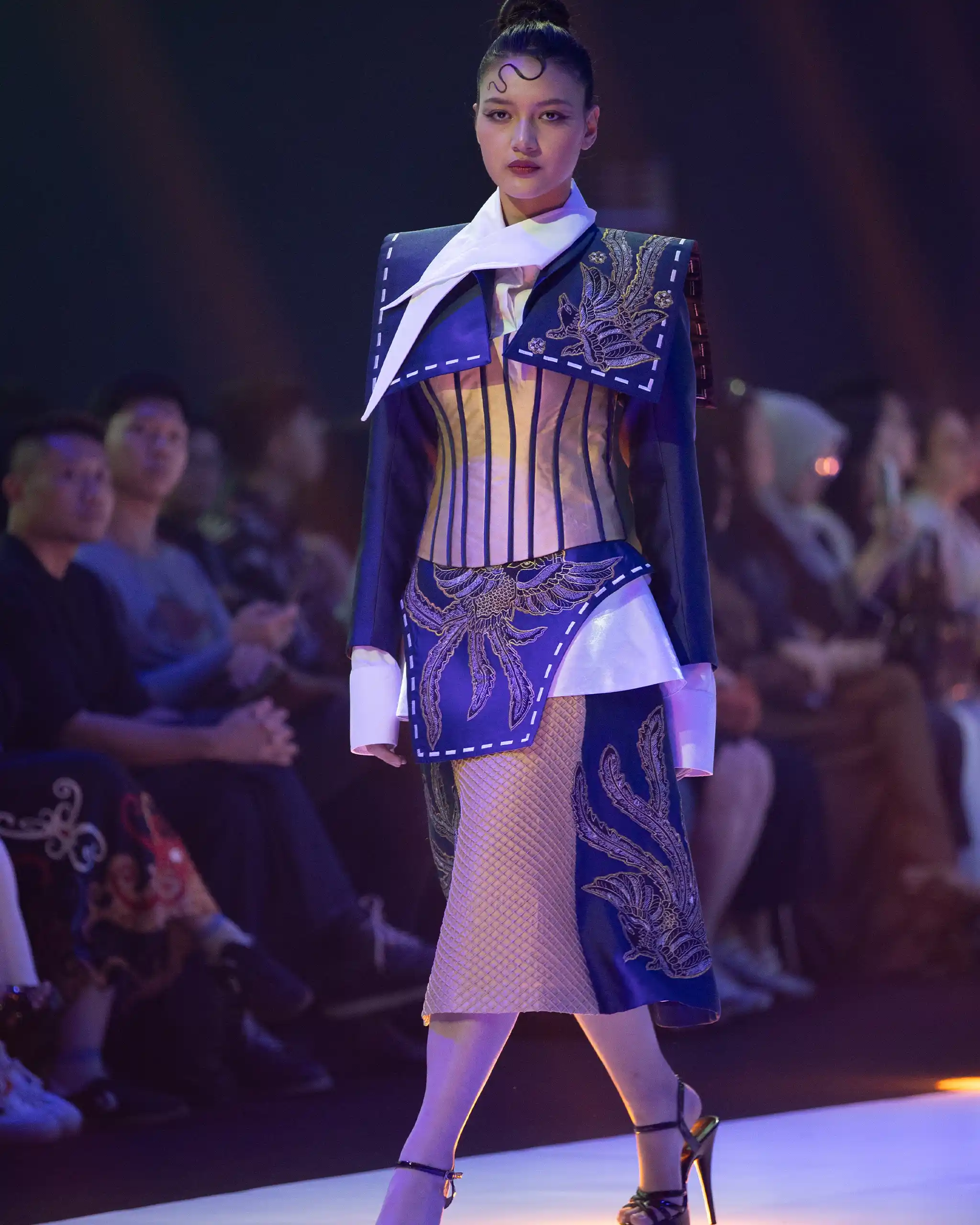 Model walks a fashion runway wearing a blue embroidered suit with a white scarf and fishnet tights, audience blurred in the background.
