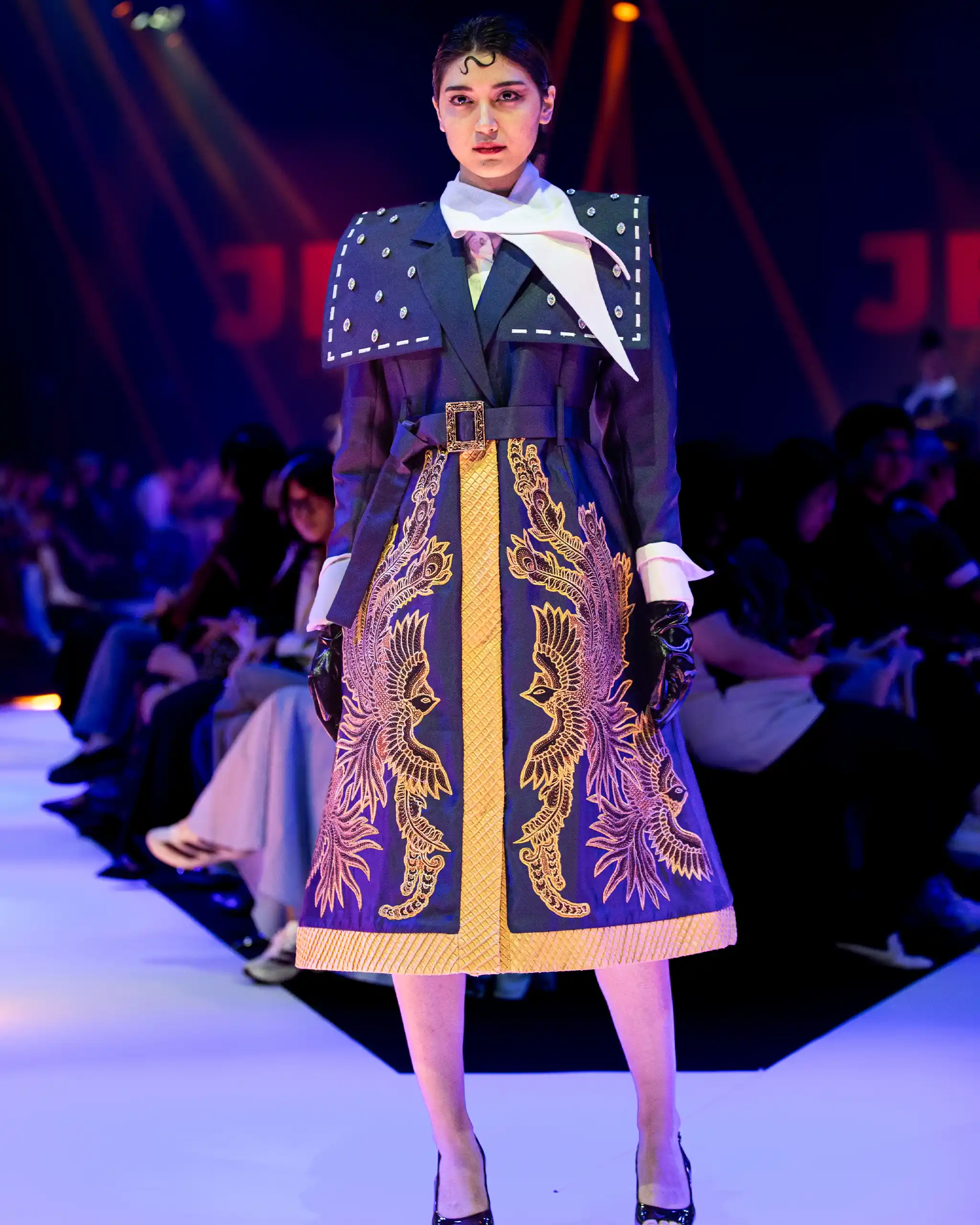 Model on a runway wearing a blue coat with gold dragon embroidery, a wide belt, and a white scarf.