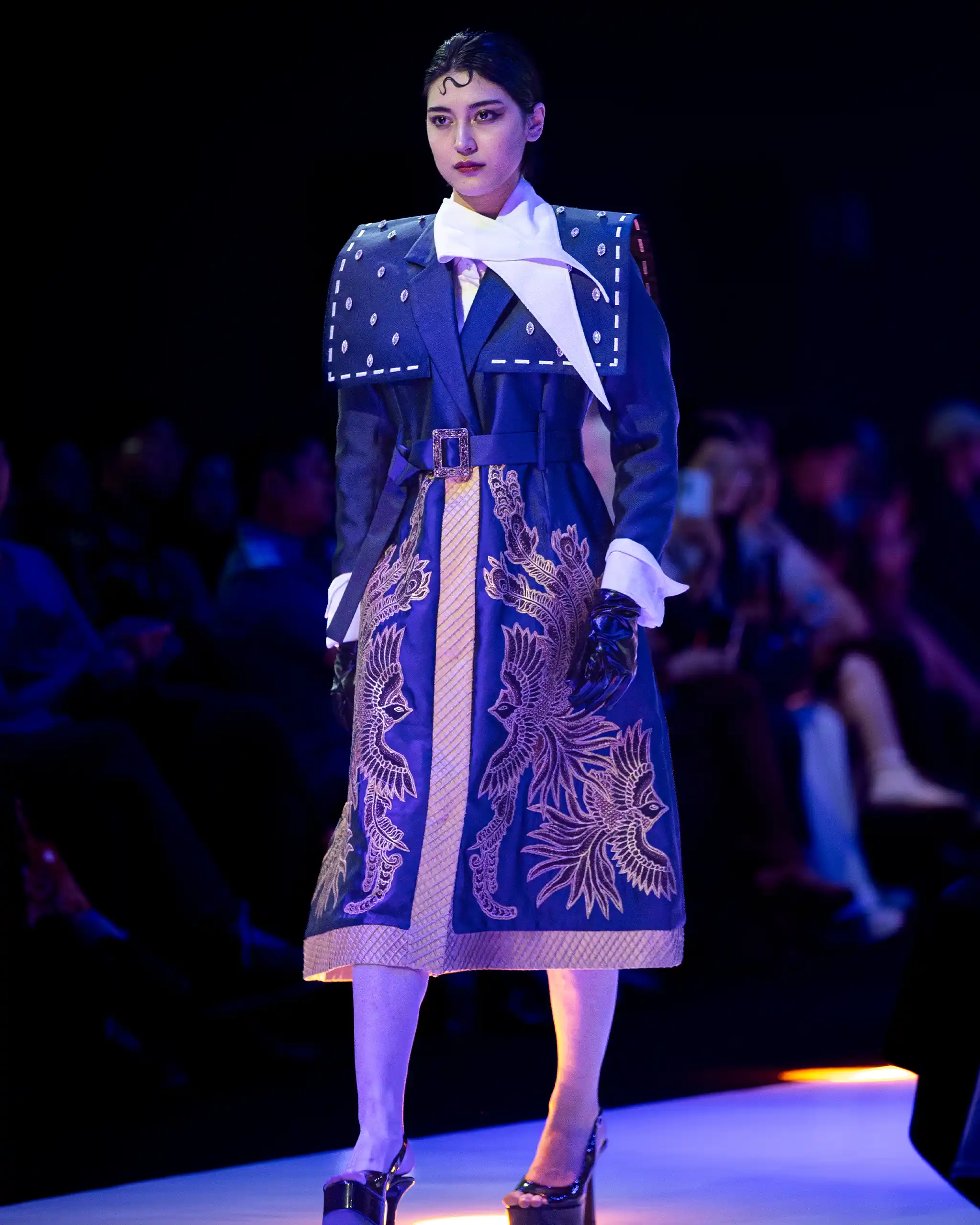 Female runway model wearing a blue cape-style coat with white scarf and dragon embroidery, standing on a lit runway.