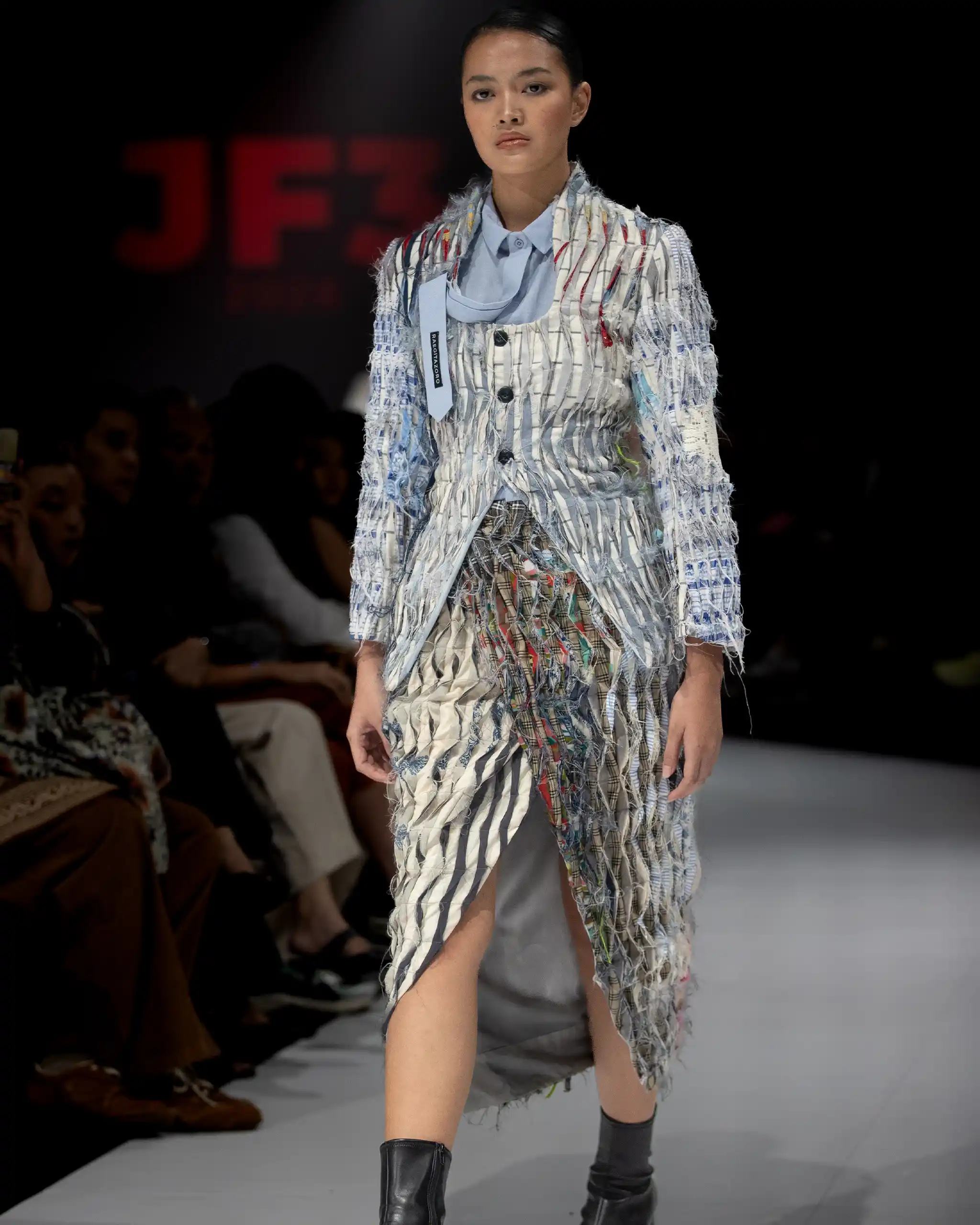 Model walks the runway in a shredded, fringe-striped jacket over a light blue button-down shirt; audience blurred in the background.