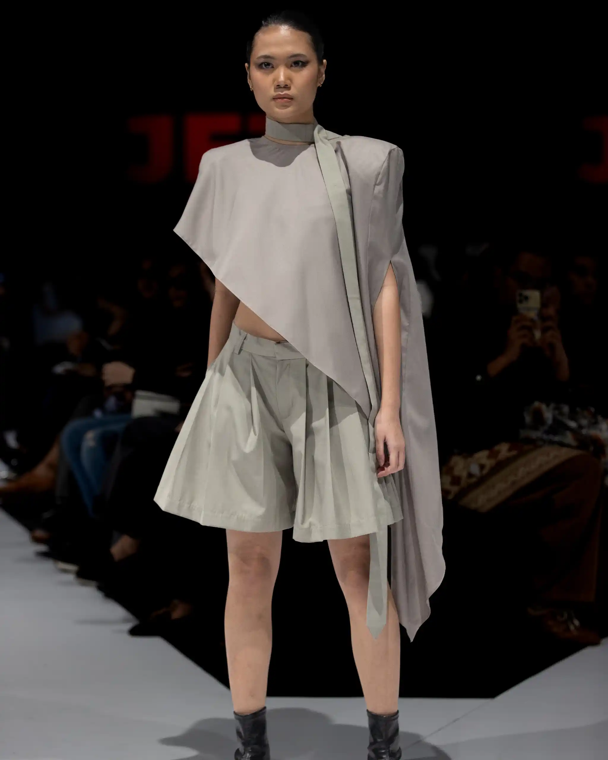 Model walking a fashion show in a light gray, asymmetrical cape blouse with matching pleated shorts or skirt; dark venue with blurred audience in background.