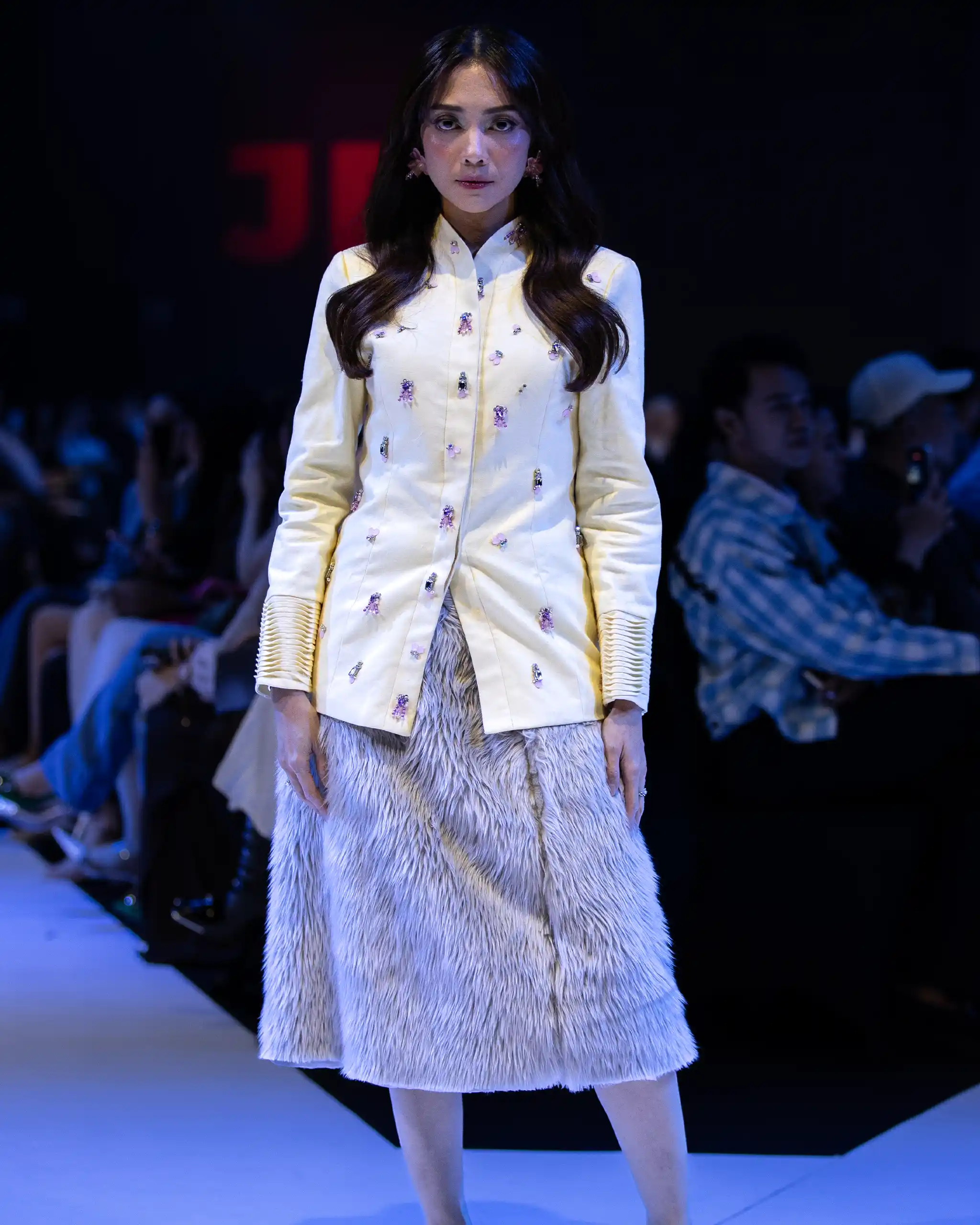 Model on runway wearing a pale yellow embroidered coat over a fur skirt, with audience in the background.