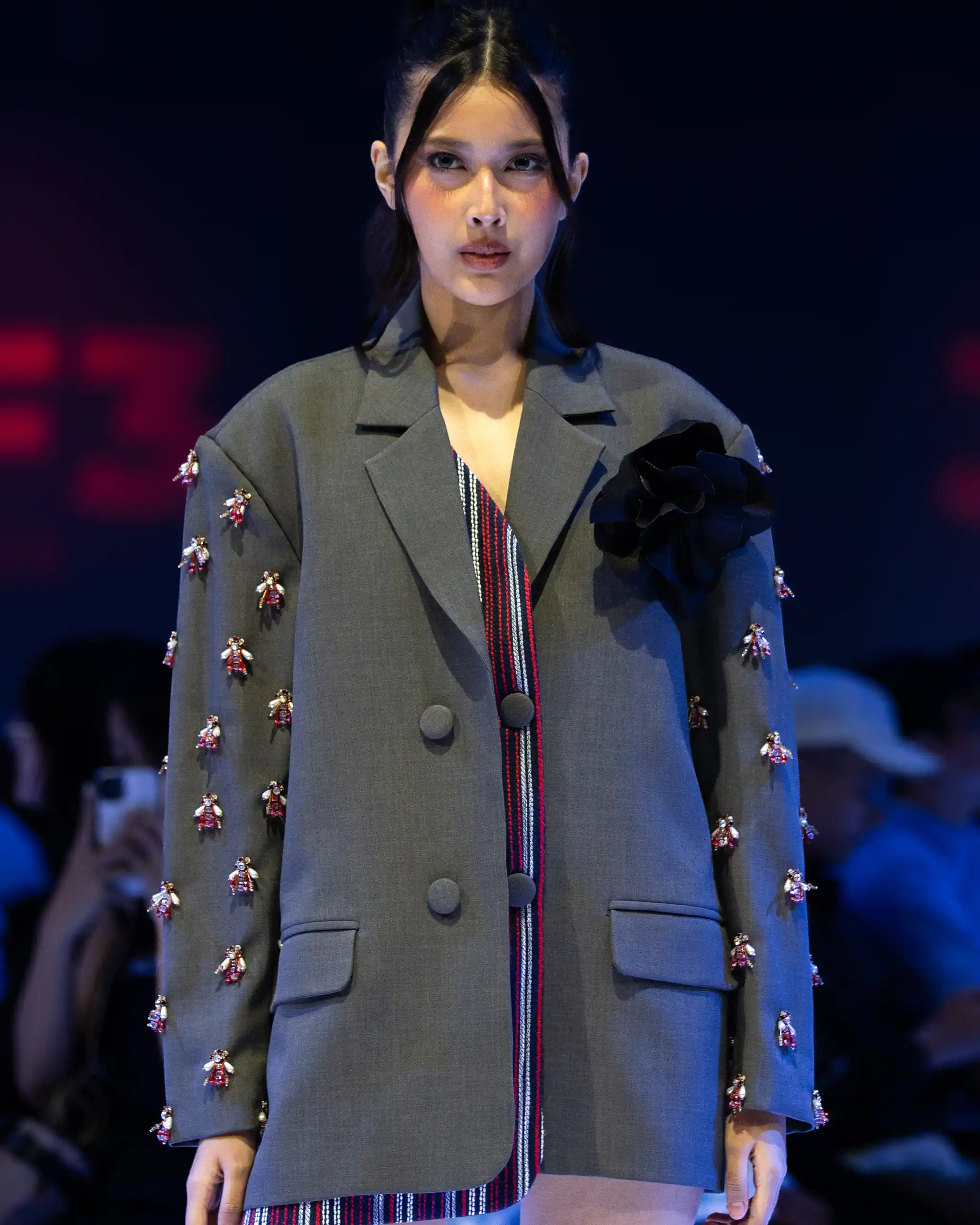 Female model in a gray blazer with floral embroidery along the sleeves, on a runway with a striped inner lining showing at the lapel.