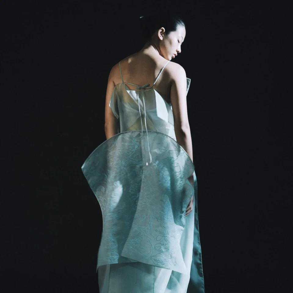Back view of a woman in a pale teal, sleeveless dress against a dark background.