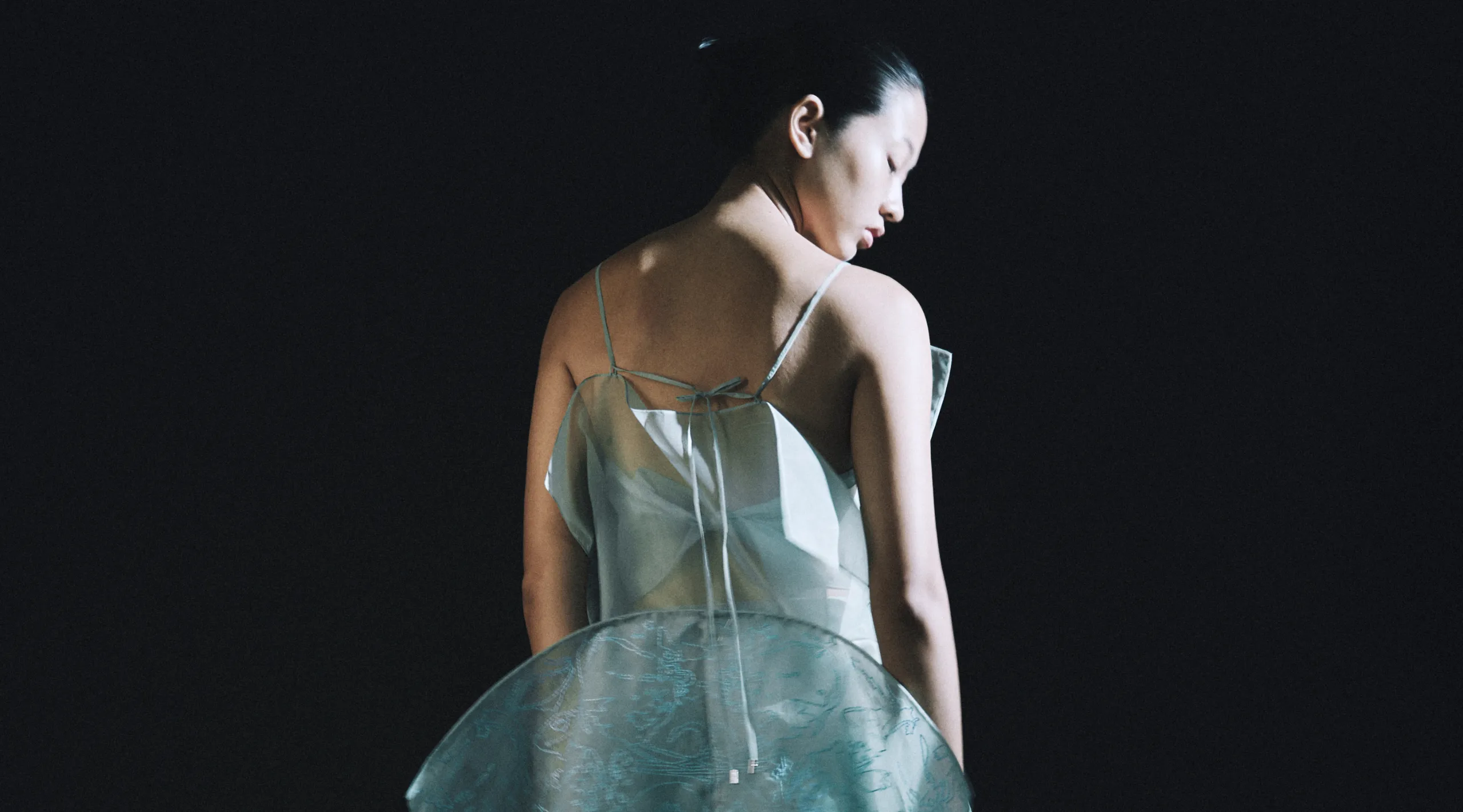 Person seen from behind wearing a pale teal, sleeveless dress with a tied back against a dark background