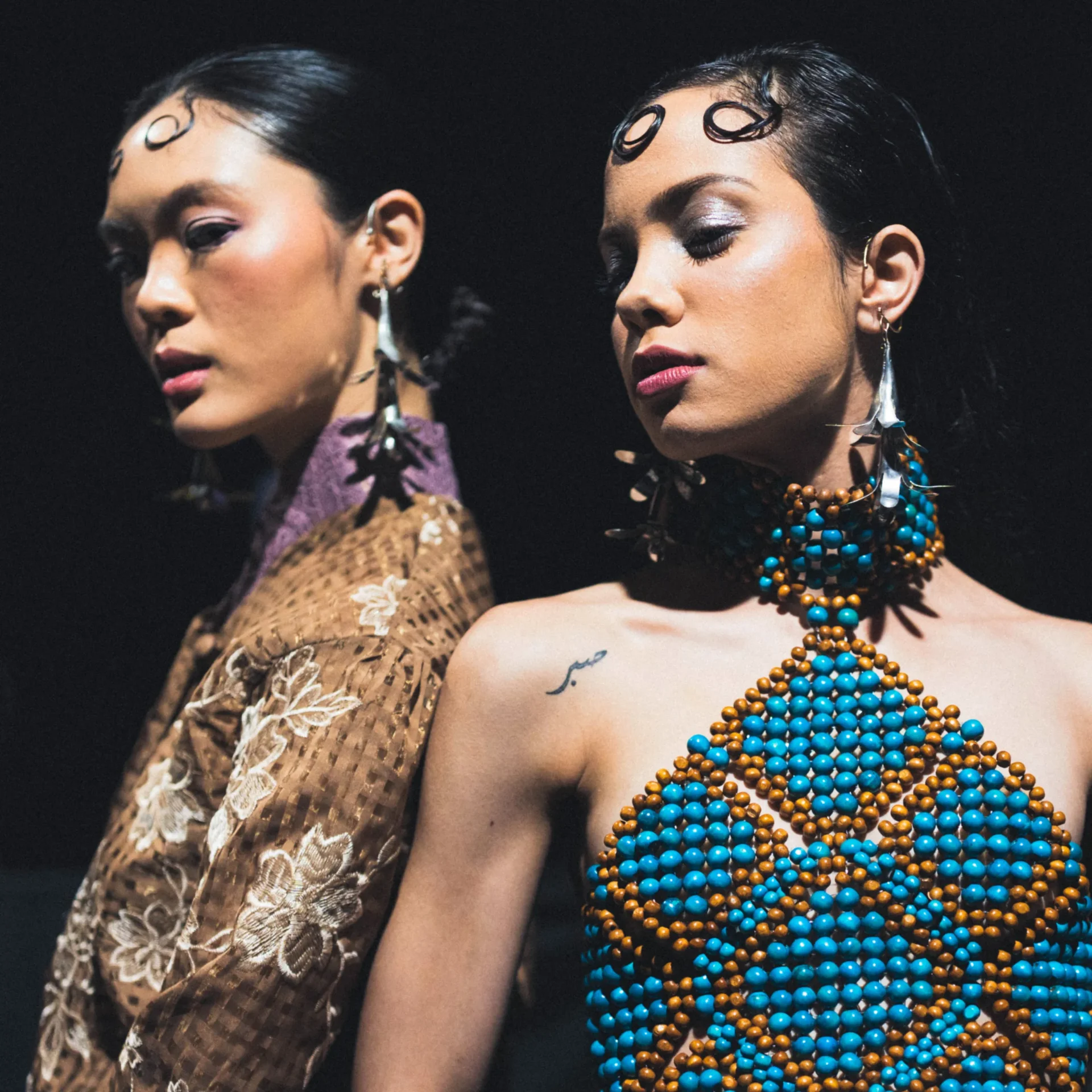 Two fashion models wear bold beaded necklaces in turquoise and orange against a dark background, close up of their shoulders and faces.