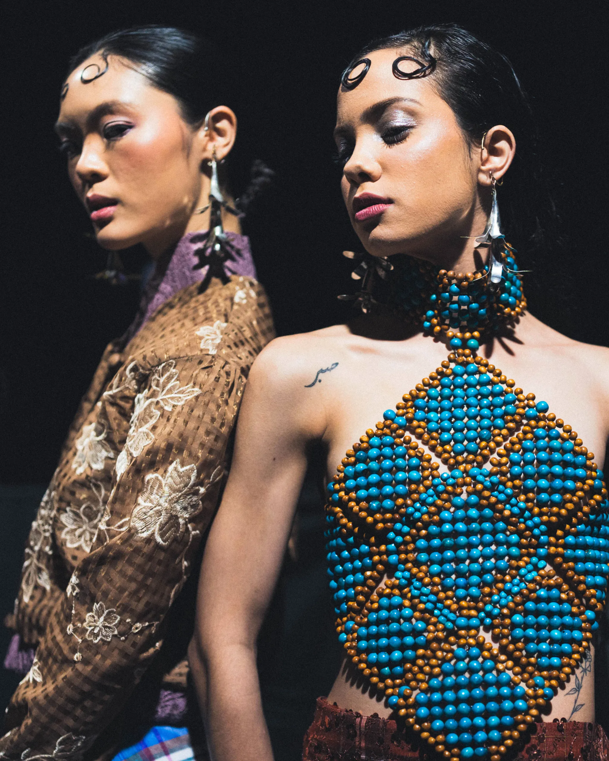 Two models pose against a dark backdrop; the right-side model wears a vibrant turquoise and brown bead necklace, while the left wears a brown floral dress.