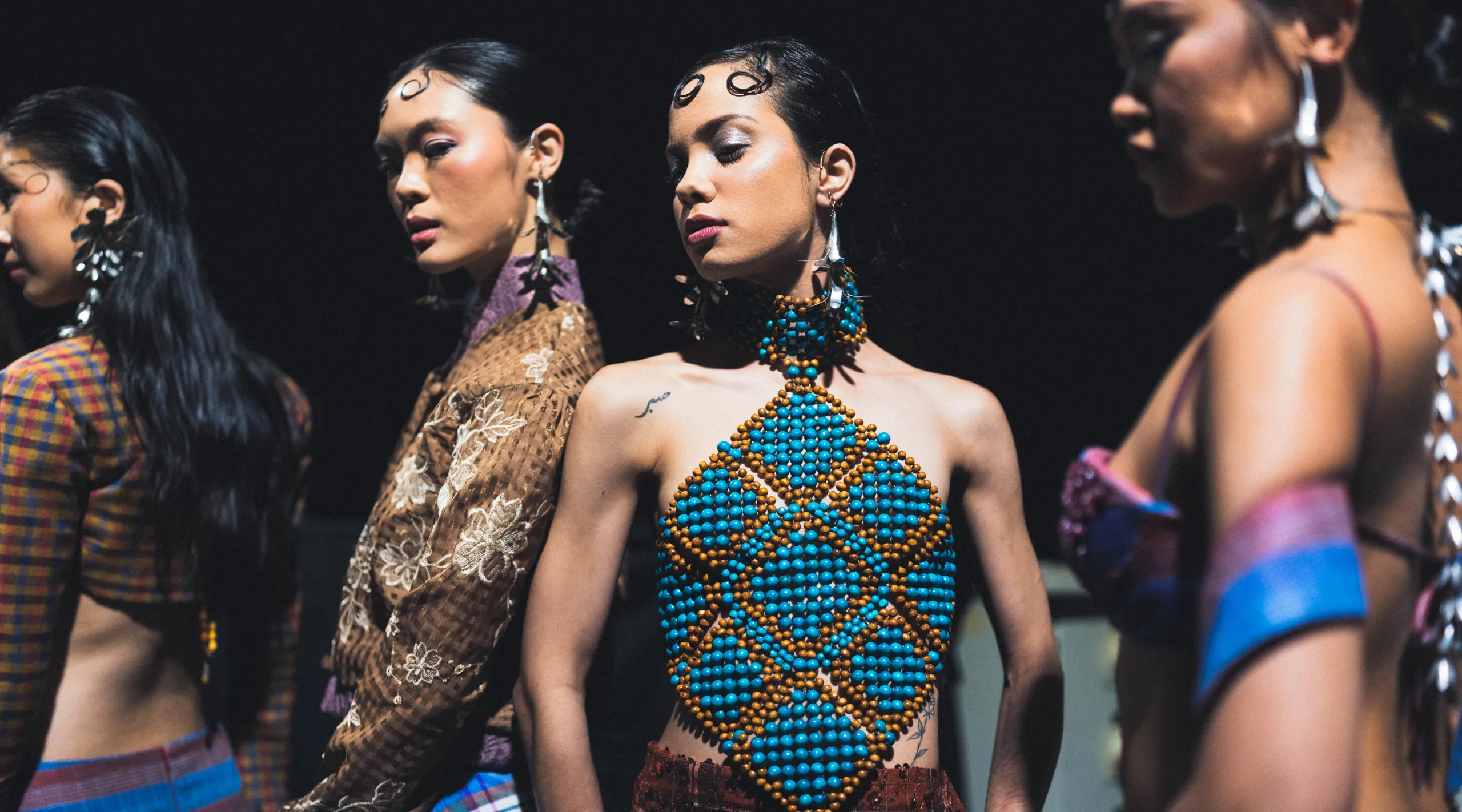 Runway models wear elaborate beadwork outfits; the center model in turquoise and orange beaded bodice stands out against a dark background.