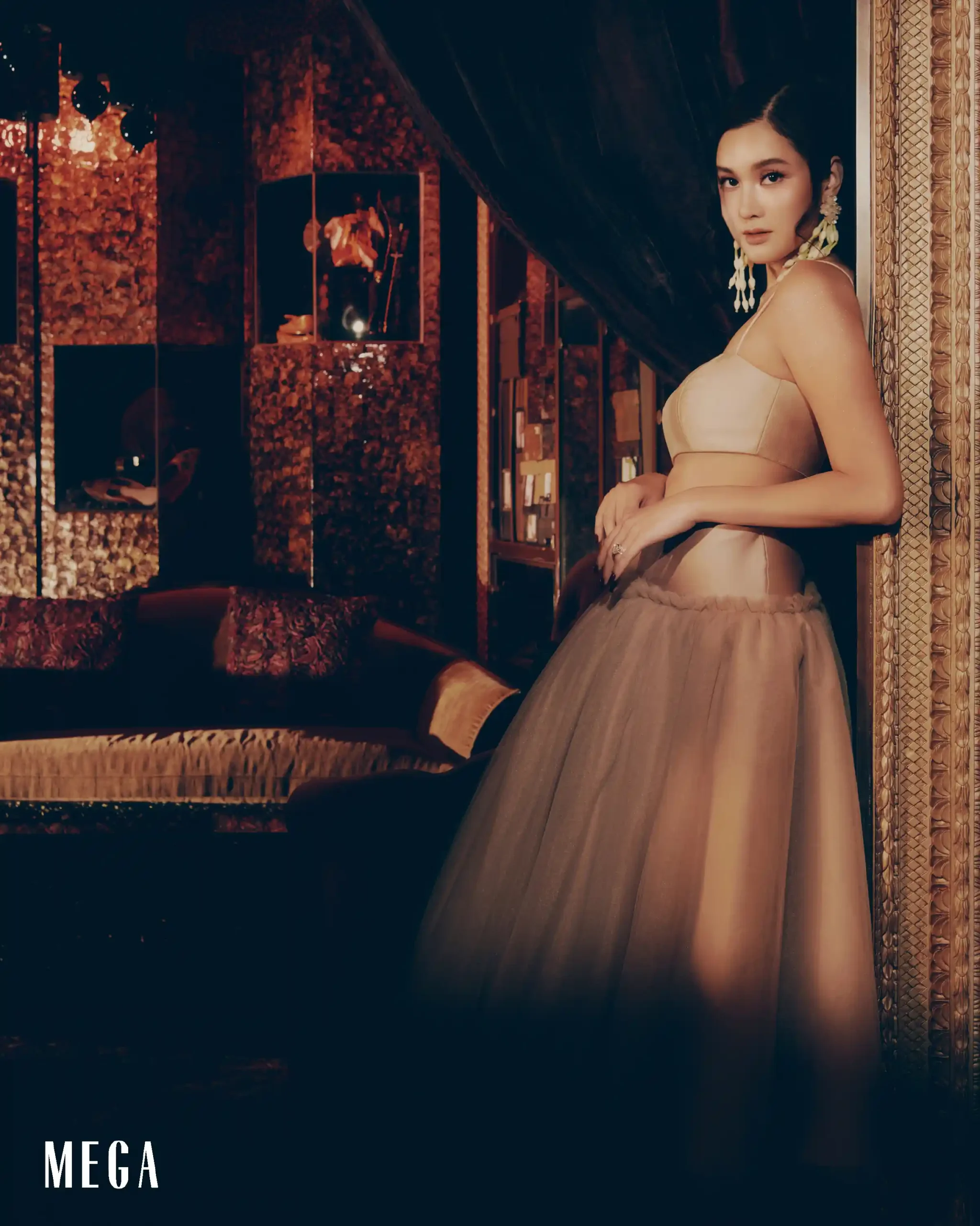 Woman in a beige gown posing against a dark, ornate wall, with 'MEGA' text visible in the bottom-left corner.