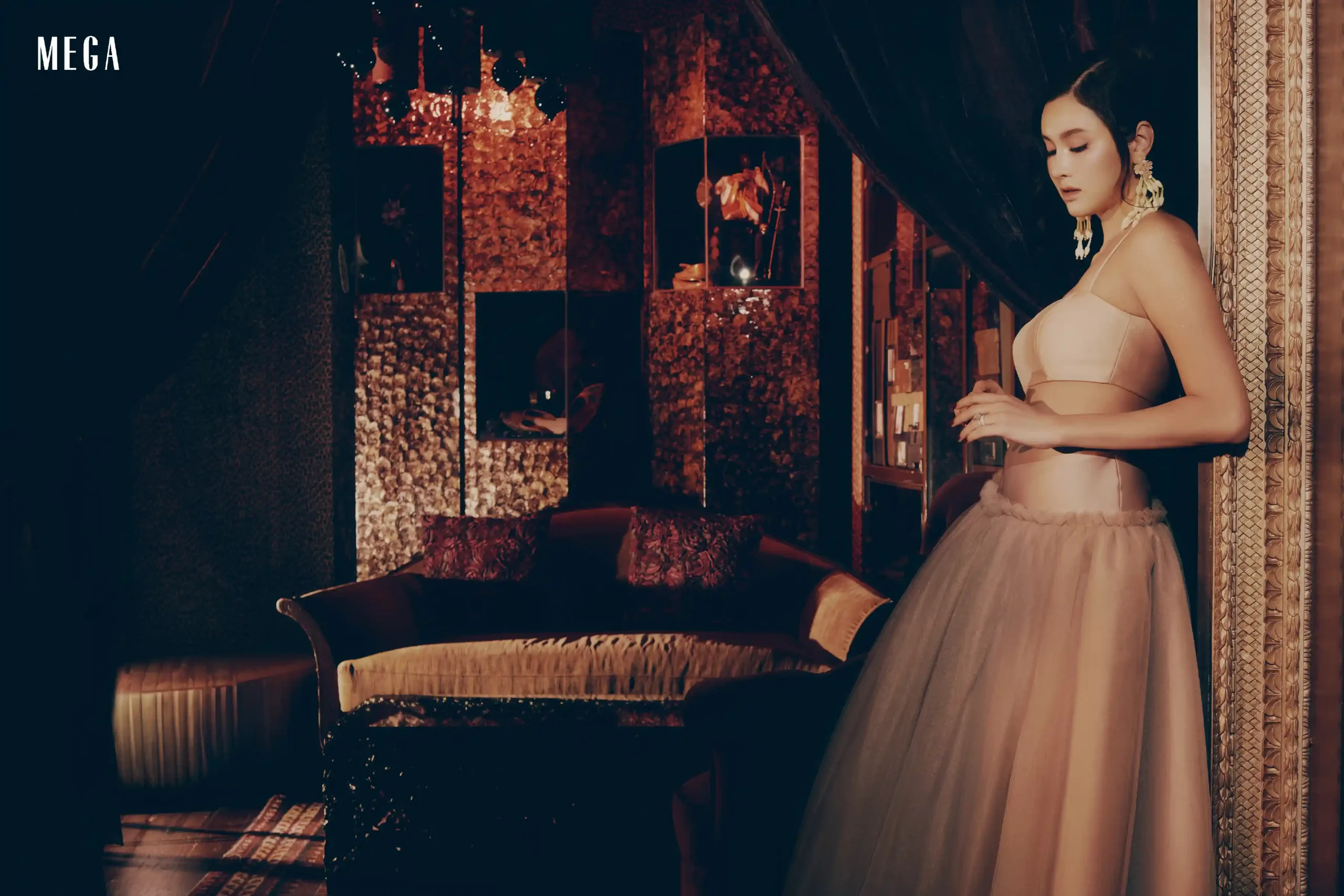 A fashionable woman in a pink strapless top and tulle skirt stands sideways in a luxurious boutique with a dark blue wall and a carved frame on the right.