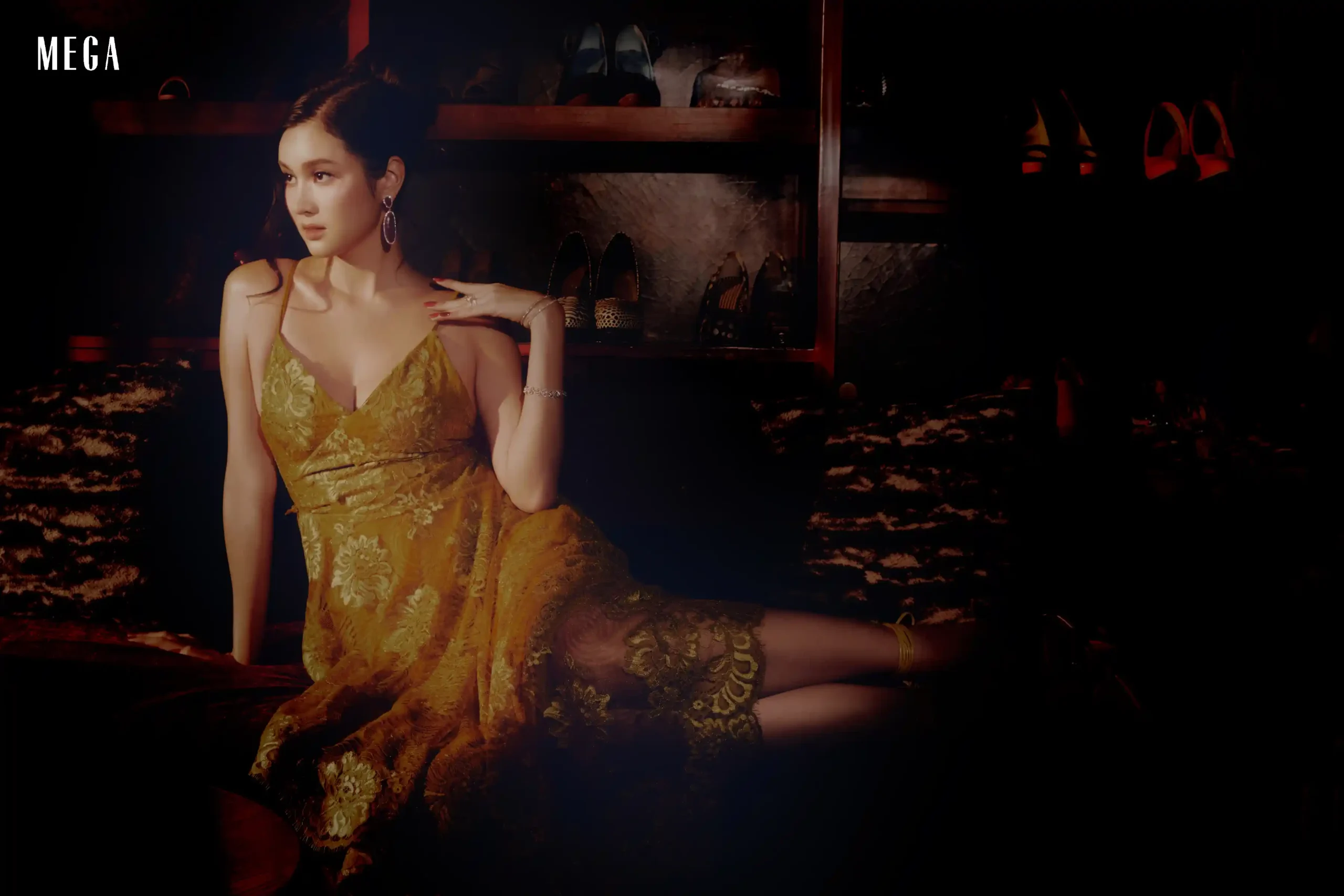 A woman in a gold lace dress sits in a dim boutique, with shelves of high-heeled shoes behind and a MEGA logo in the top-left corner.
