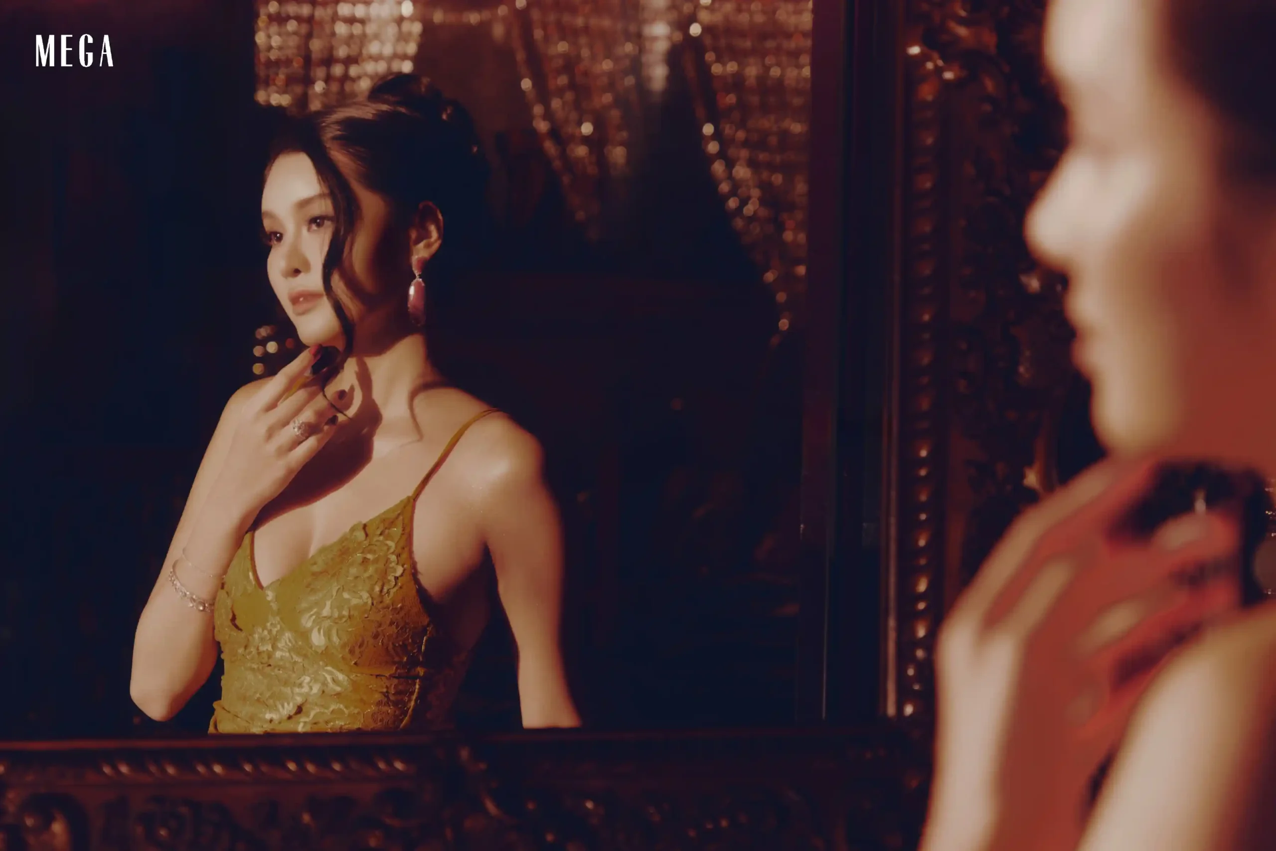 Woman in a green lace dress touching her neck, gazing at her reflection in a dark, warmly lit room with city lights outside.