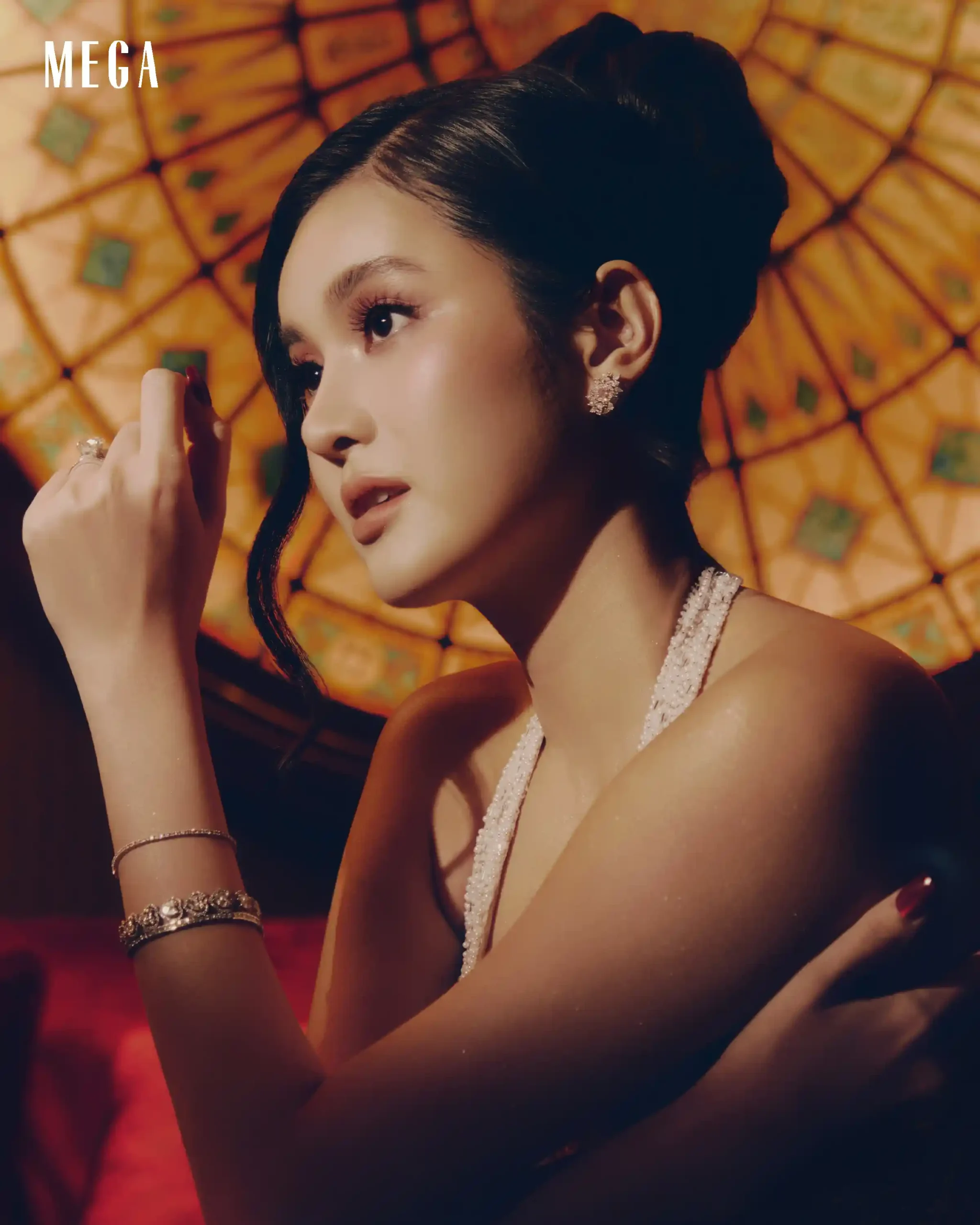 Close-up of a woman wearing diamond earrings and a pearl necklace, turned to the left in warm lighting with an orange patterned background.