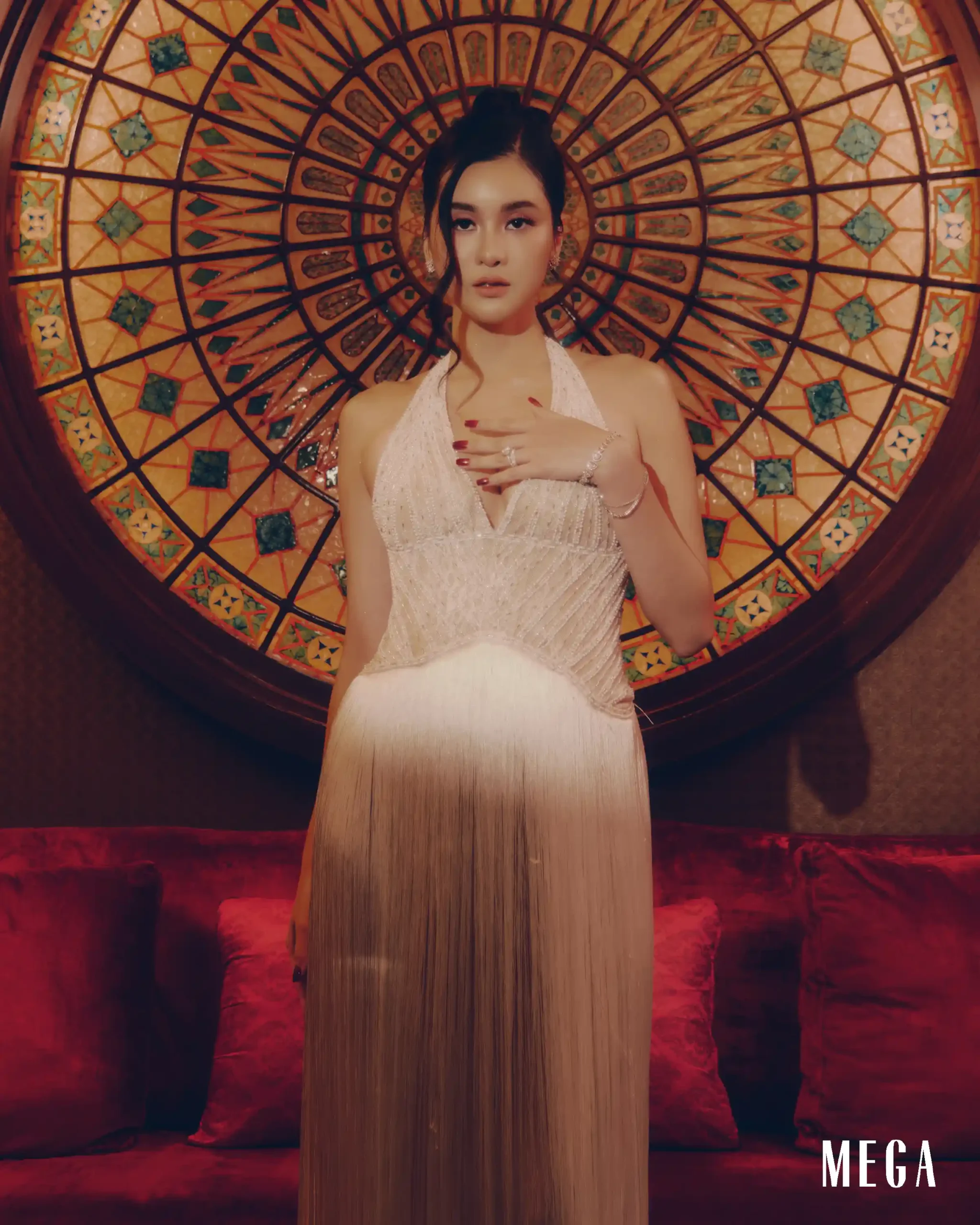 A woman in a shimmering white gown stands before a large circular stained-glass wall, hands crossed at chest, with red nails and jewelry visible, MEGA watermark at bottom right.