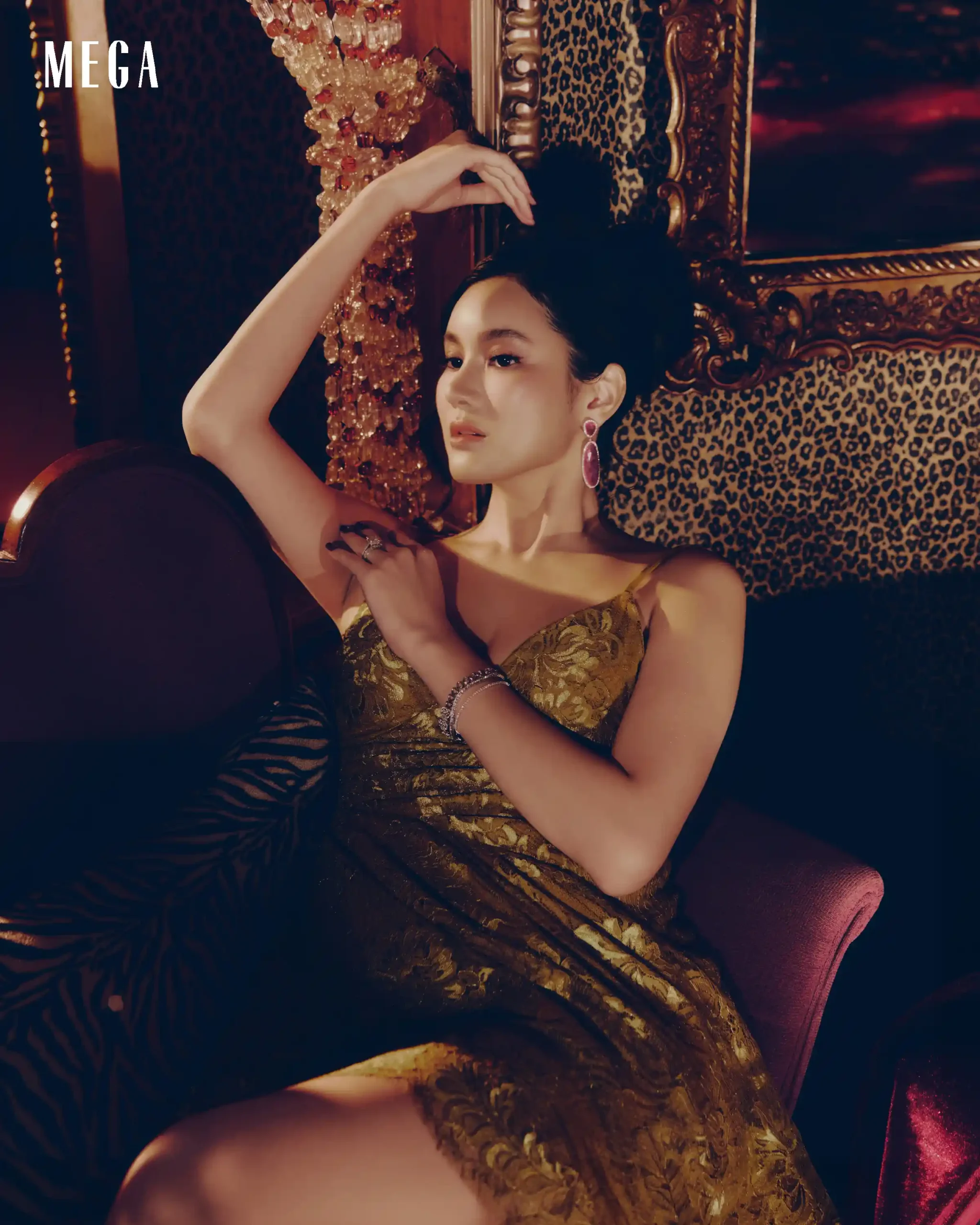 Woman in a gold patterned dress lounging on an ornate carved chair with leopard upholstery and pearl earrings.